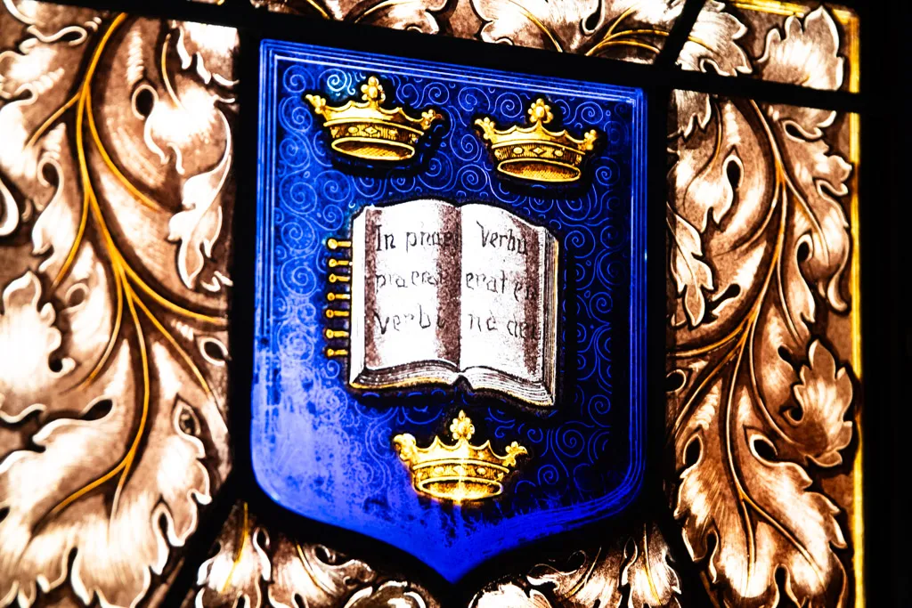 Stained glass window depicting a blue shield with three golden crowns and an open book with Latin text in the center, surrounded by ornate gold and brown floral patterns.