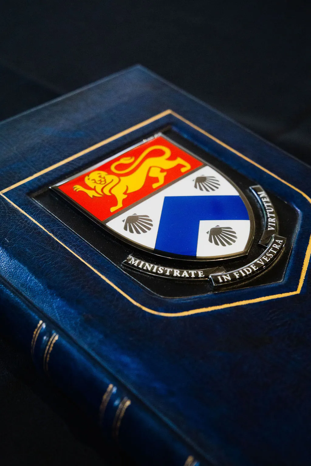 Close-up of a dark blue book cover with a coat of arms featuring a yellow lion on red and blue shields, labeled 'MINISTRATE IN FIDE VESTRA VIRTUTEM'.