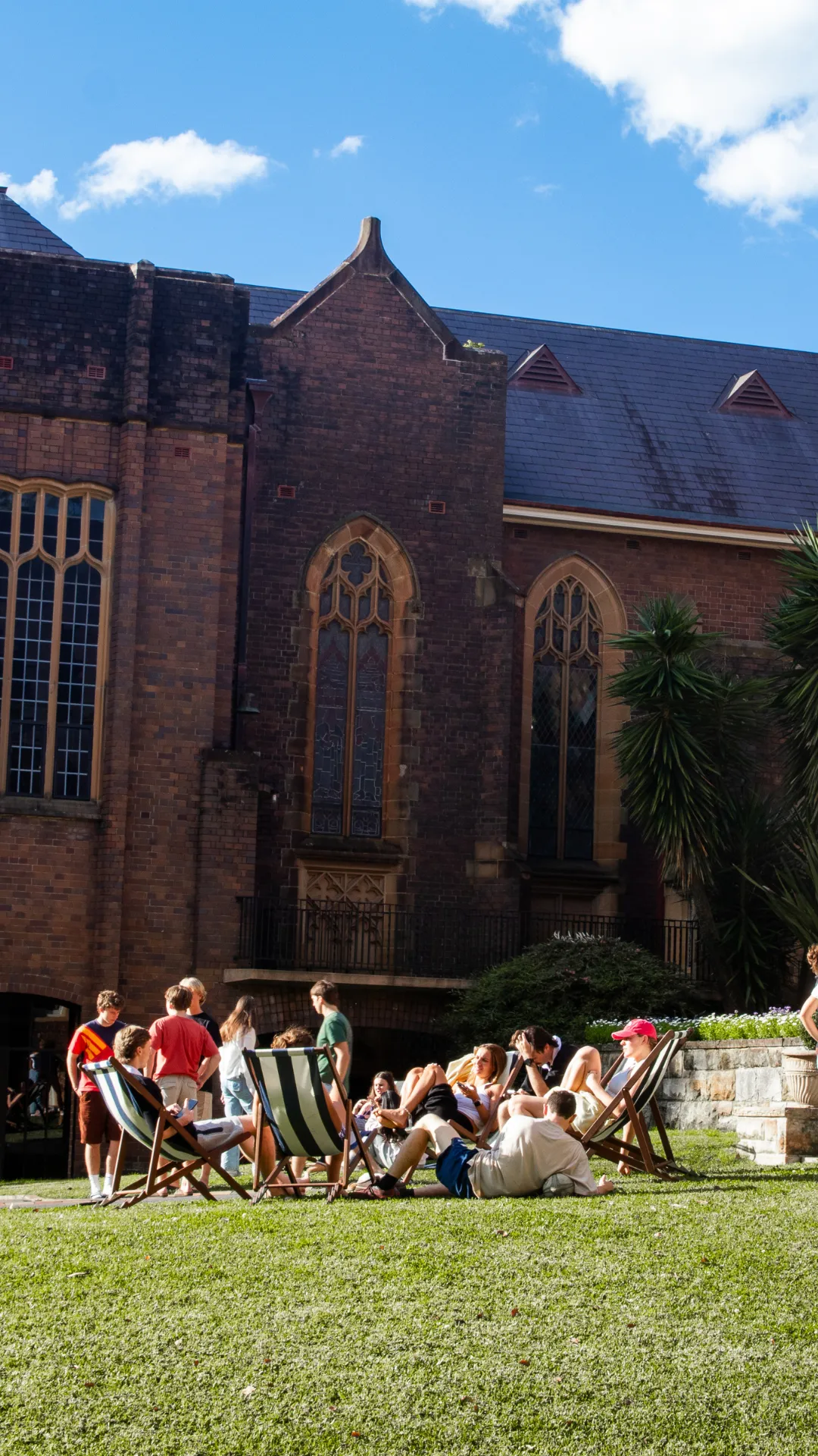 Group of young people relaxing on deck chairs and sitting on grass outside a brick building with large stained glass windows on a sunny day.