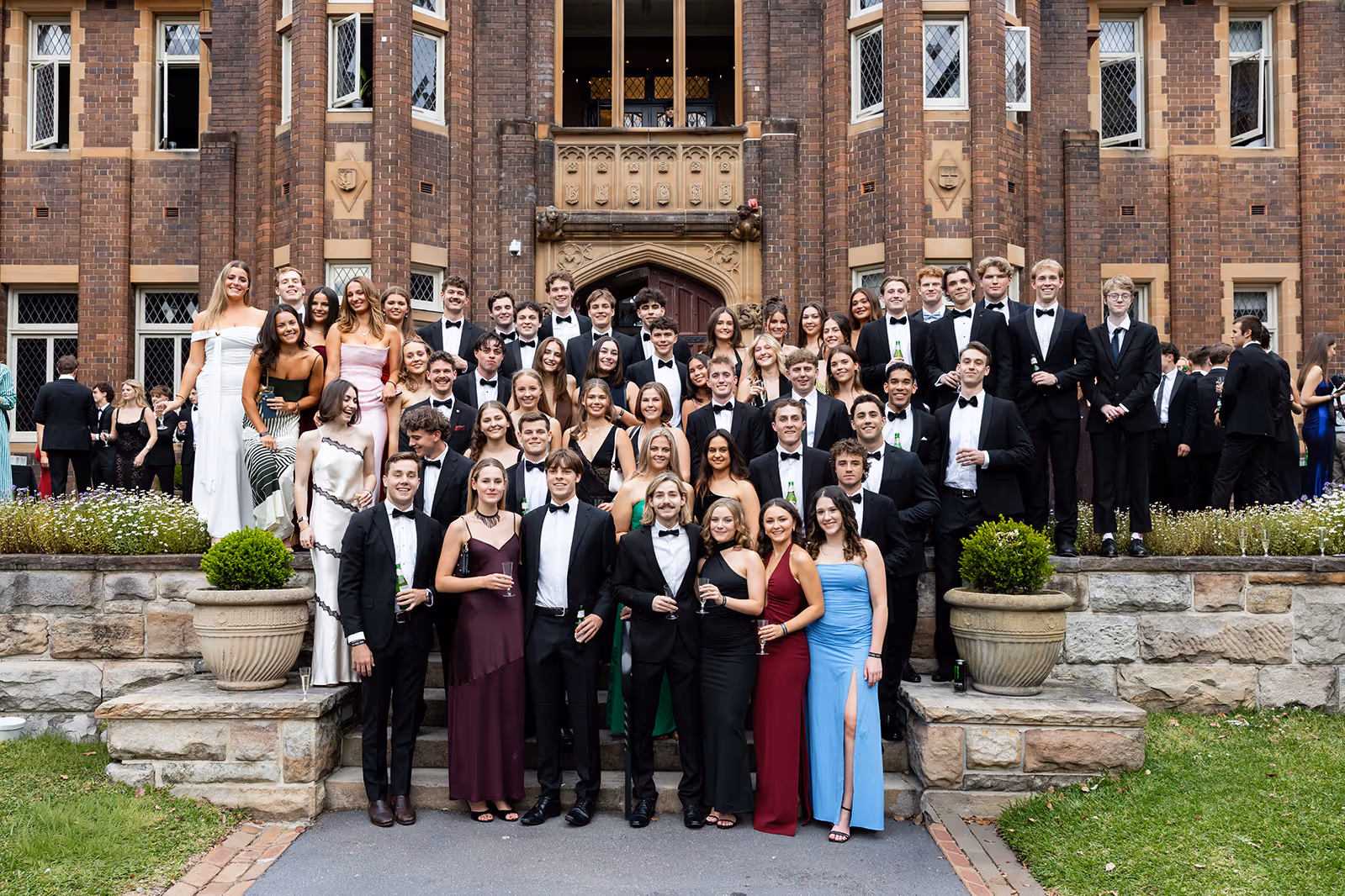 Large group of young adults dressed in formal evening wear posing for a group photo outside a brick building with decorative windows.