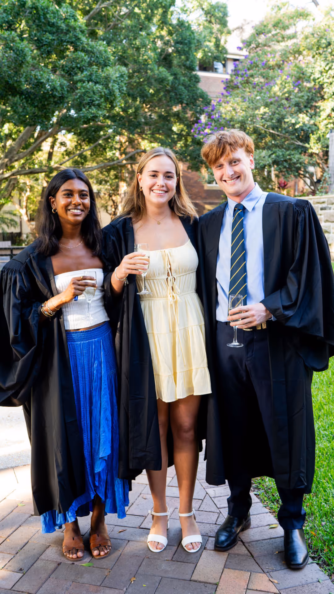 Three graduates in robes smiling outdoors, holding champagne glasses, with trees and a building in the background.