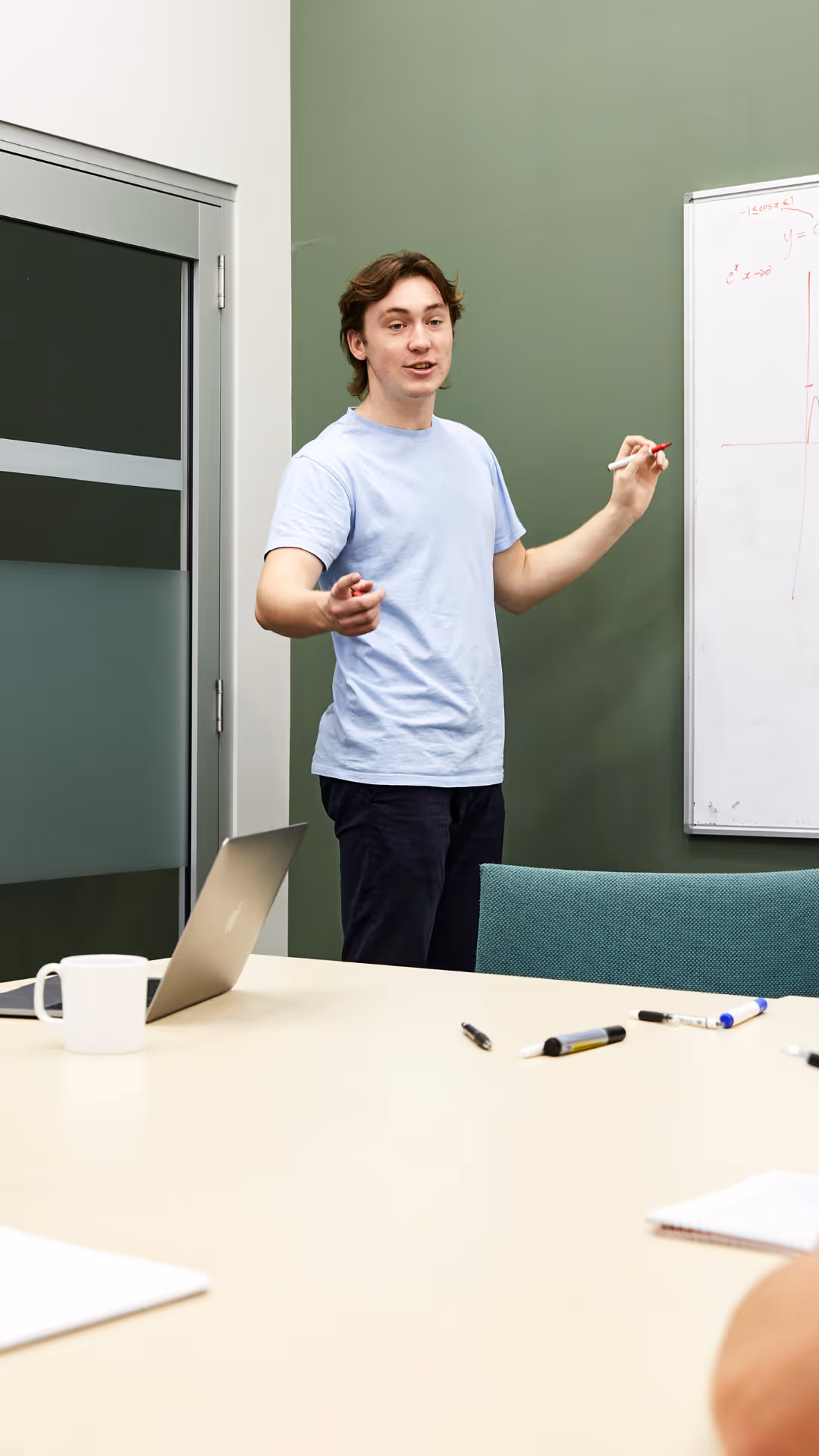 Young man in a light blue shirt explaining math on a whiteboard in a classroom.