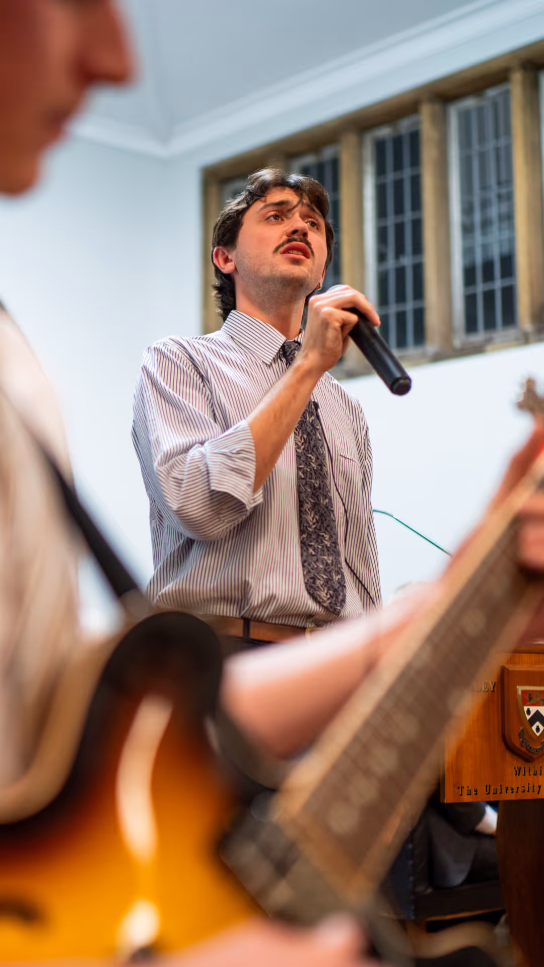 Man in a striped shirt and patterned tie singing into a microphone inside a building with large windows.