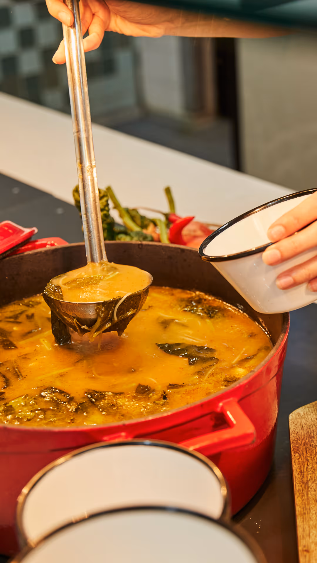 Hand ladling soup with greens from a red pot into a white bowl with black rim.
