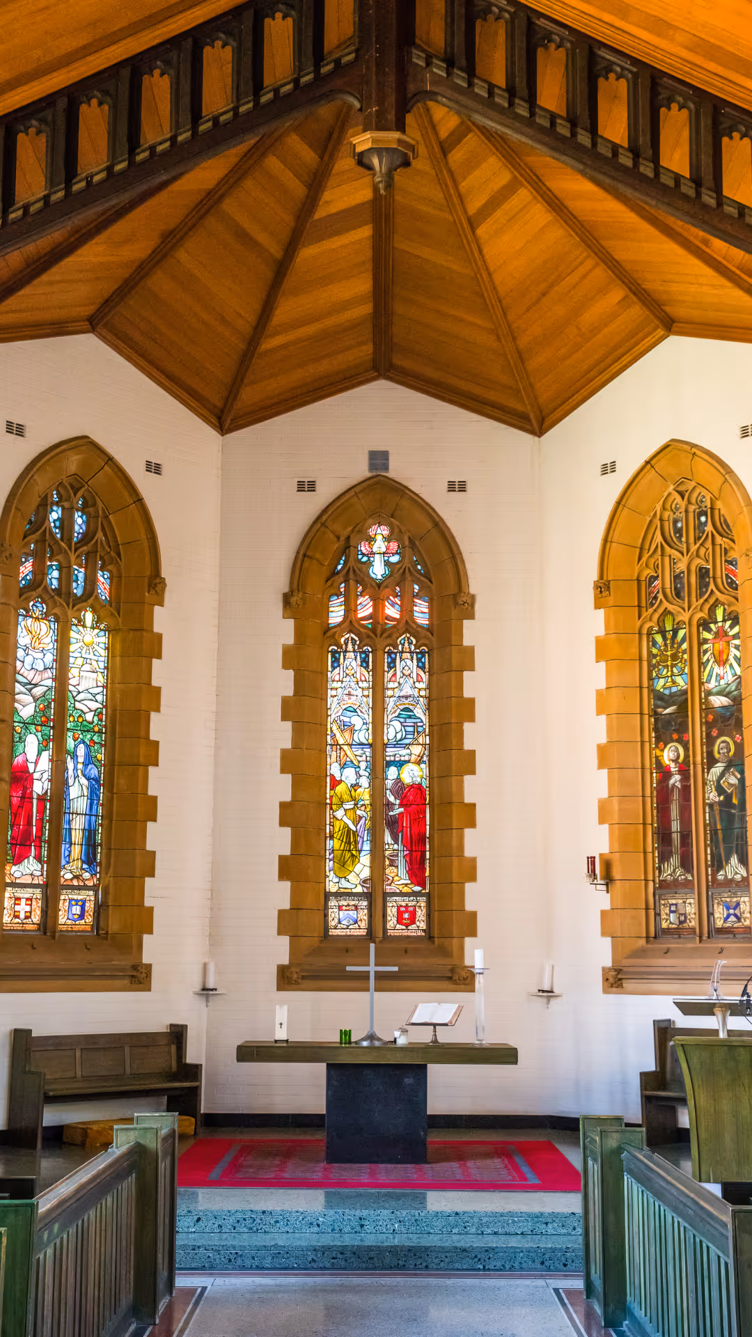 Interior of a church altar featuring three stained glass windows depicting religious figures, a central wooden altar with a cross, candles, and an open Bible, all under a wooden vaulted ceiling.