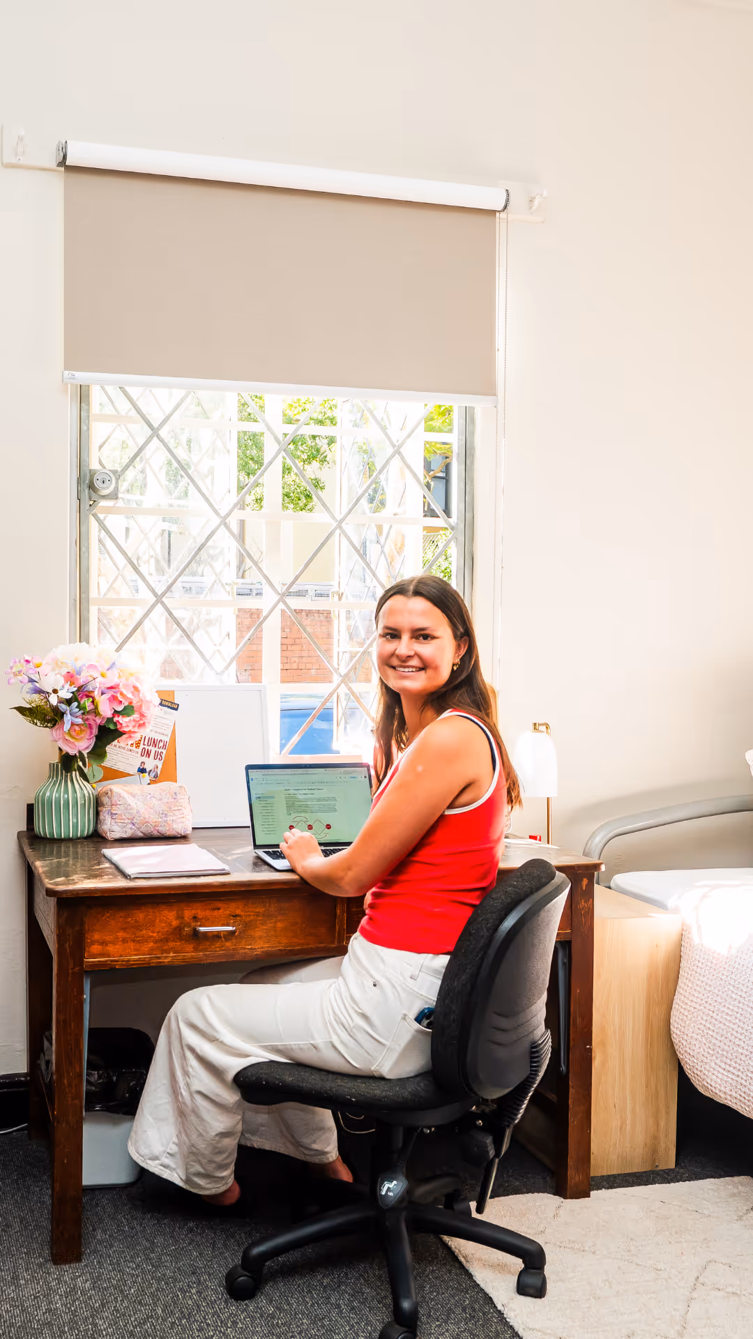 Smiling woman in red top and white pants sitting on office chair at wooden desk with laptop by a window with blinds.