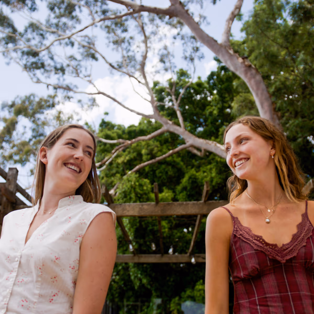 Two smiling women looking at each other outdoors with green trees and a wooden pergola in the background.