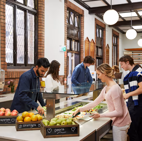 Students serving themselves fresh fruit and salad from a cafeteria buffet in a bright dining hall.