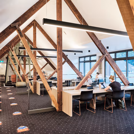 Modern study space with exposed wooden beams and several people working at desks near large windows.