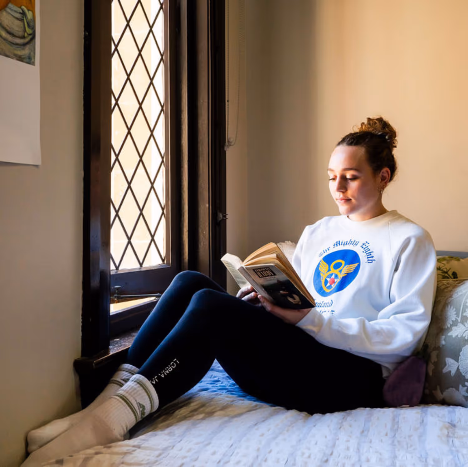 Young woman sitting on a bed by a window reading a book, wearing a white sweatshirt and dark leggings.