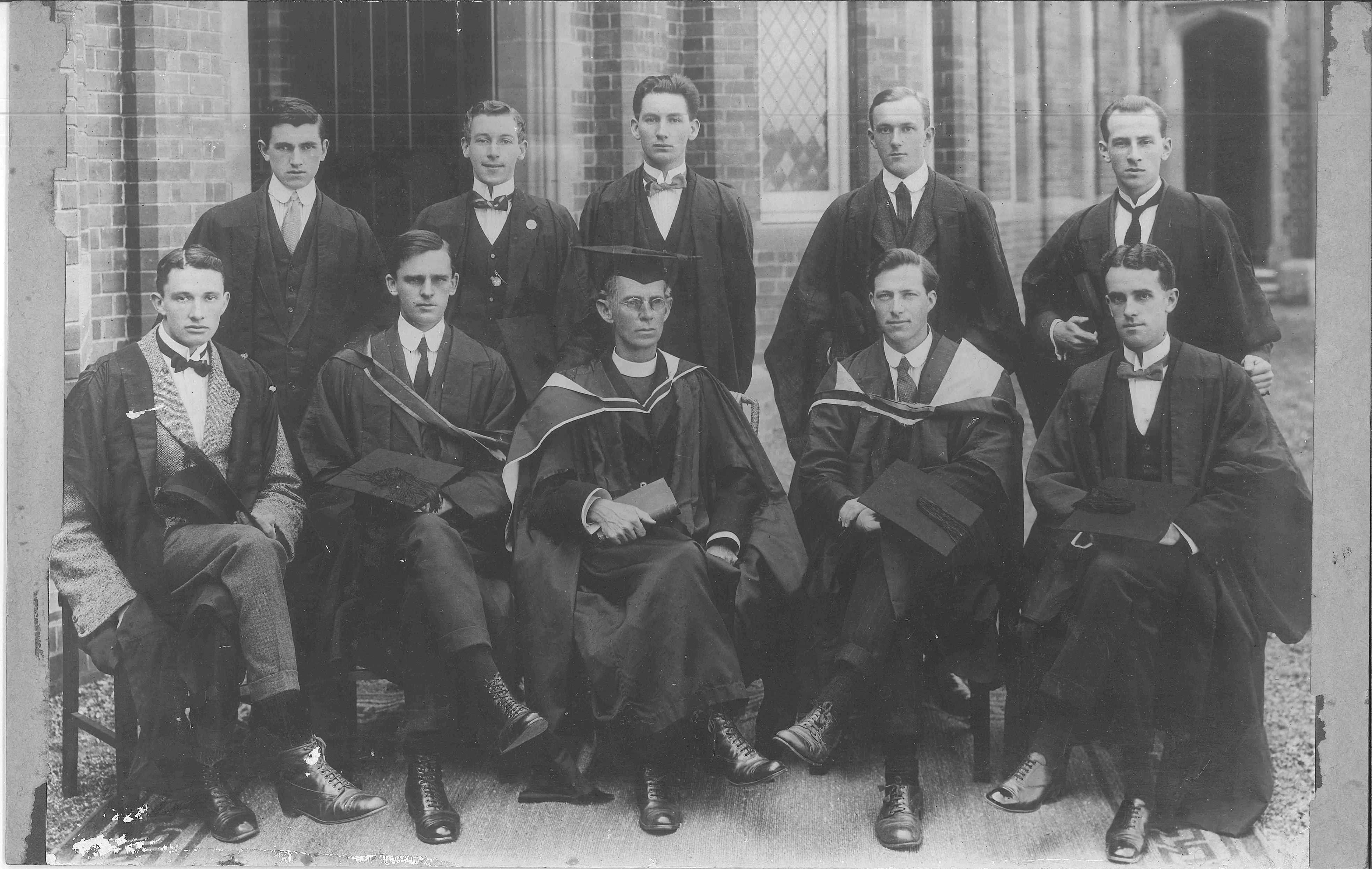 Black and white photo of eleven young men in academic gowns and formal attire, seated and standing in front of a brick building with large windows.