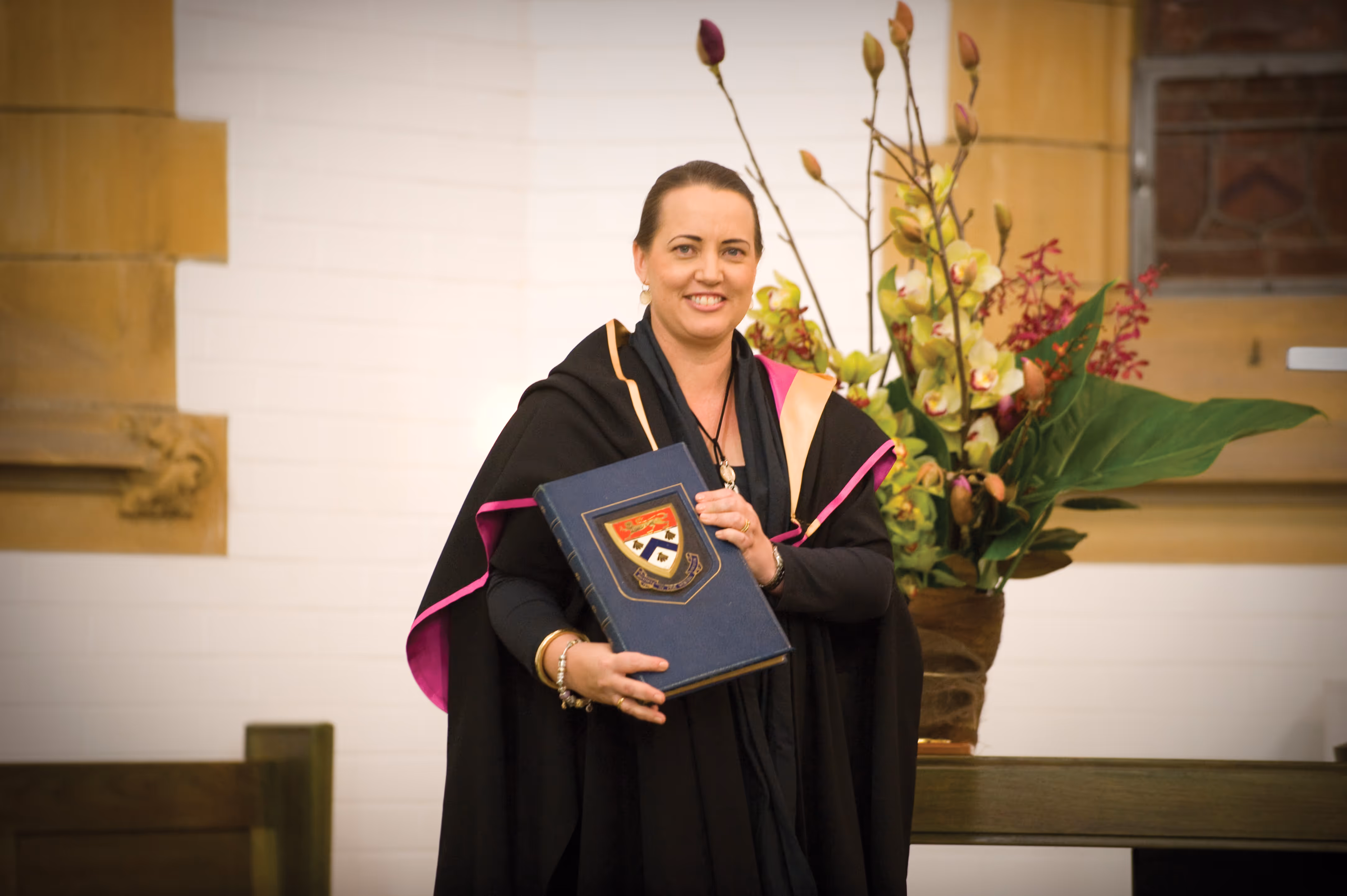 Smiling woman in graduation gown holding a large blue book with a crest, standing indoors next to a vase with orchids and green leaves.