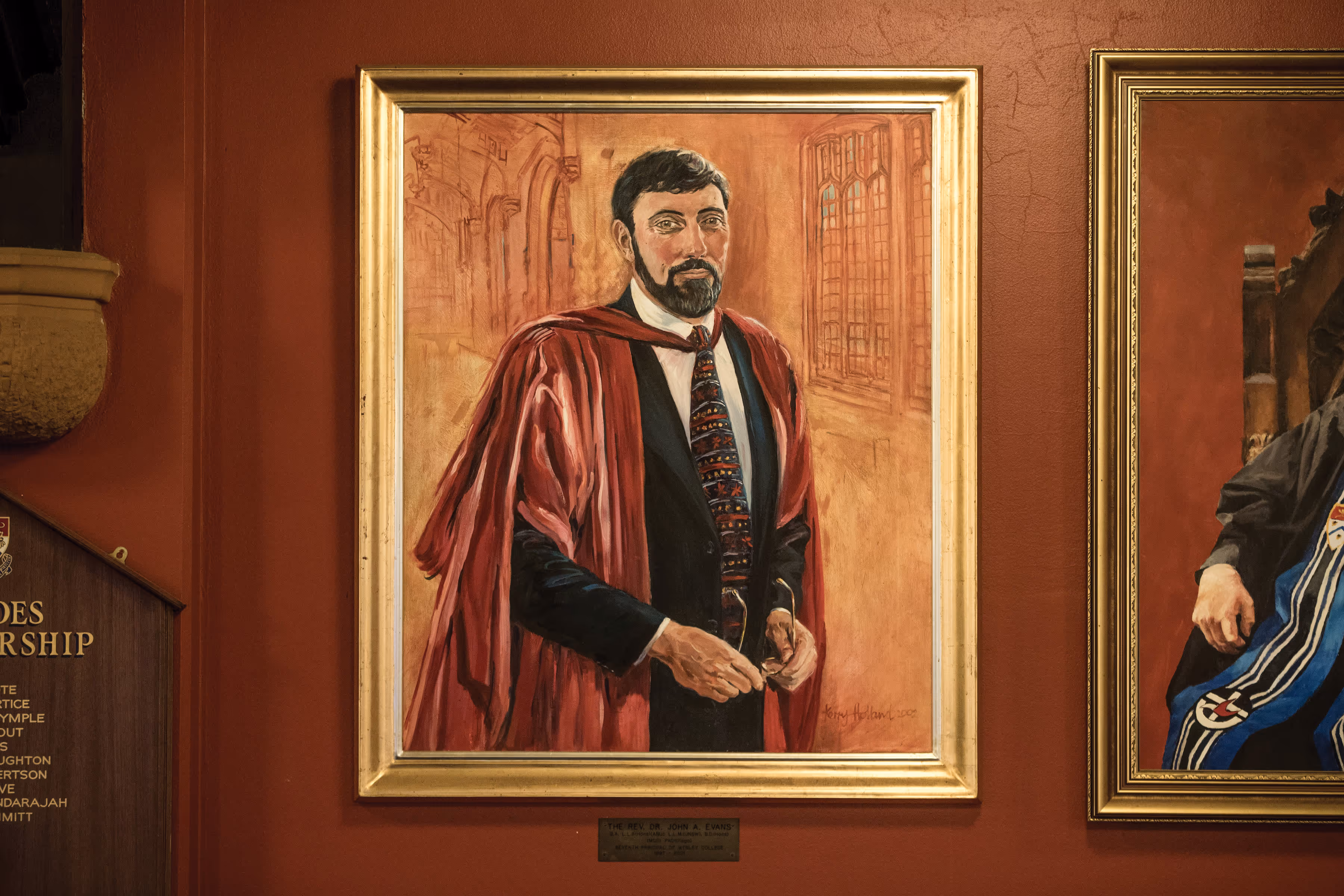 Framed portrait of a man in academic gown and colorful tie, standing indoors with arched windows in the background.
