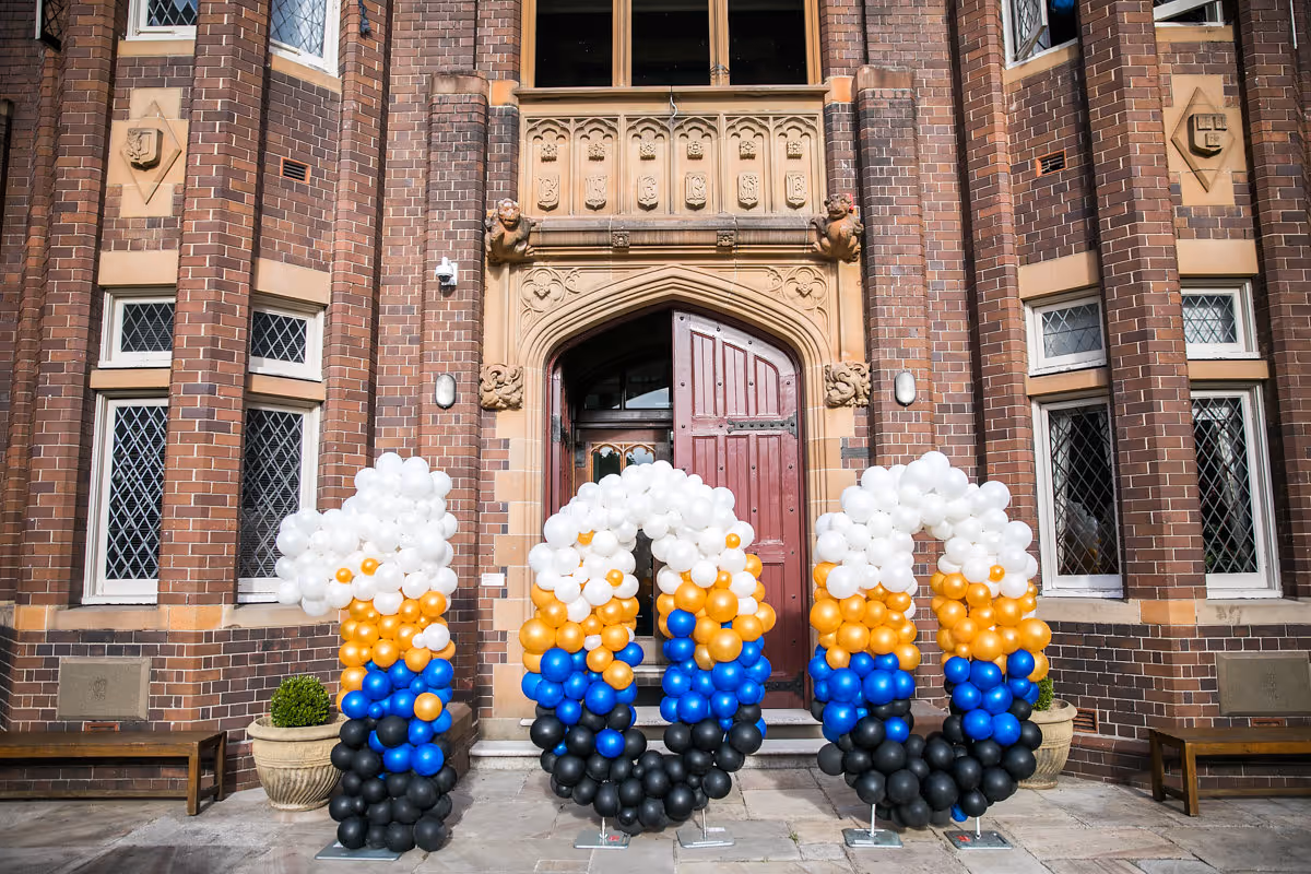 Three balloon arrangements in white, gold, blue, and black form the numbers 100 in front of a brick building with a large red door.