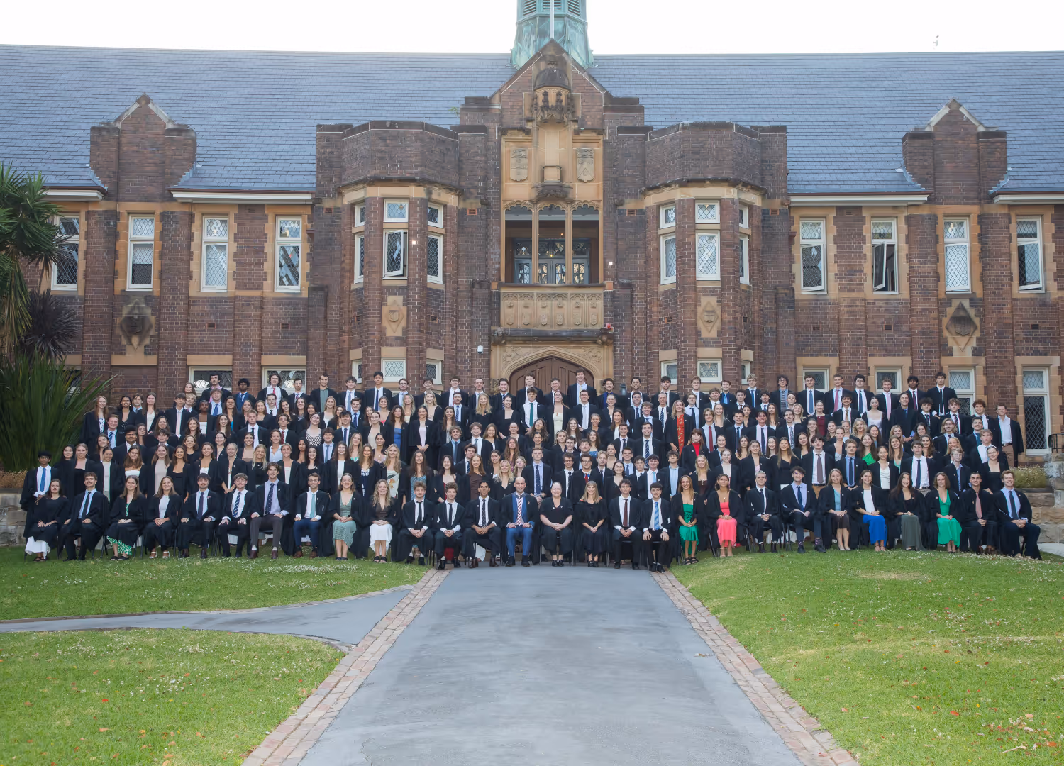 Large group of formally dressed students and staff posing for a college group photo in front of a historic brick academic building.