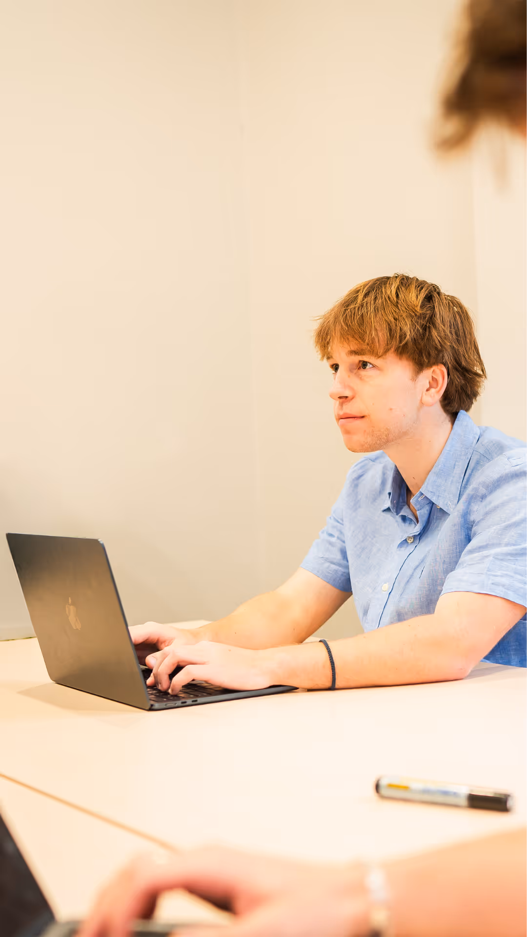 Young man with auburn hair wearing a blue shirt works on a laptop at a light-colored table in a neutral room.