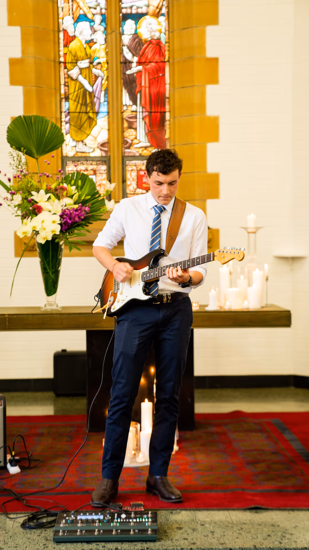 Young man in white shirt and striped tie playing electric guitar in a church with lit candles and stained glass window behind him.