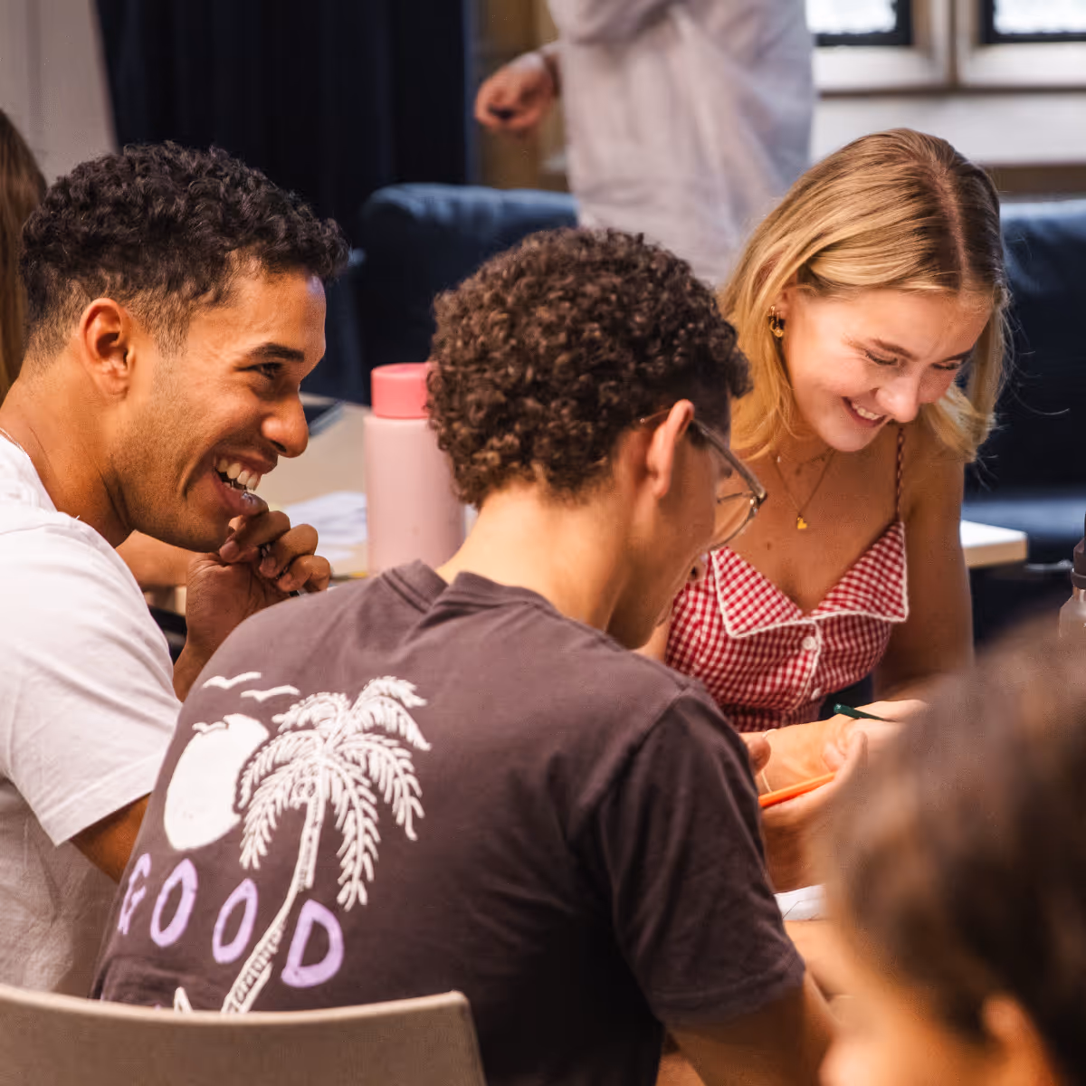 Three young adults smiling and engaging together at a table, one wearing a black shirt with a palm tree design.