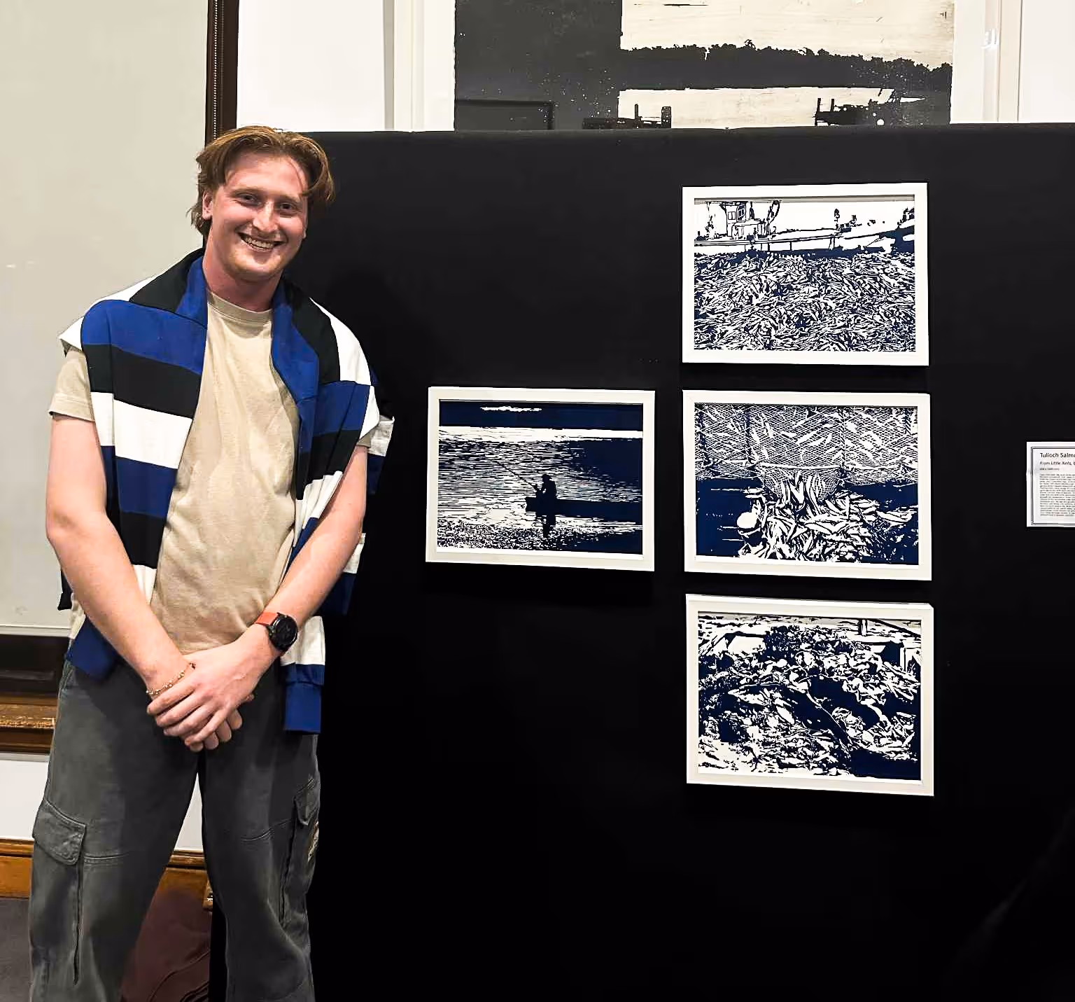 Smiling young man with a blue, black, and white striped sweater draped over his shoulders standing beside four framed blue-and-white prints mounted on a black display board.