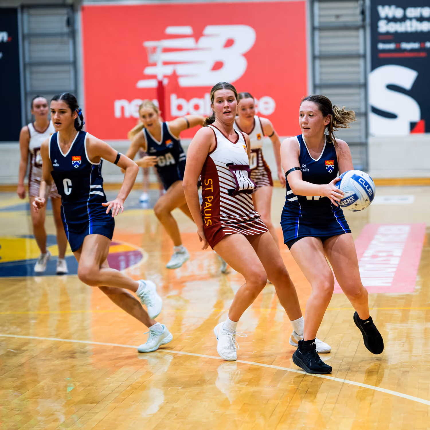 Young women playing indoor netball, one holding the ball while others move defensively on the court.