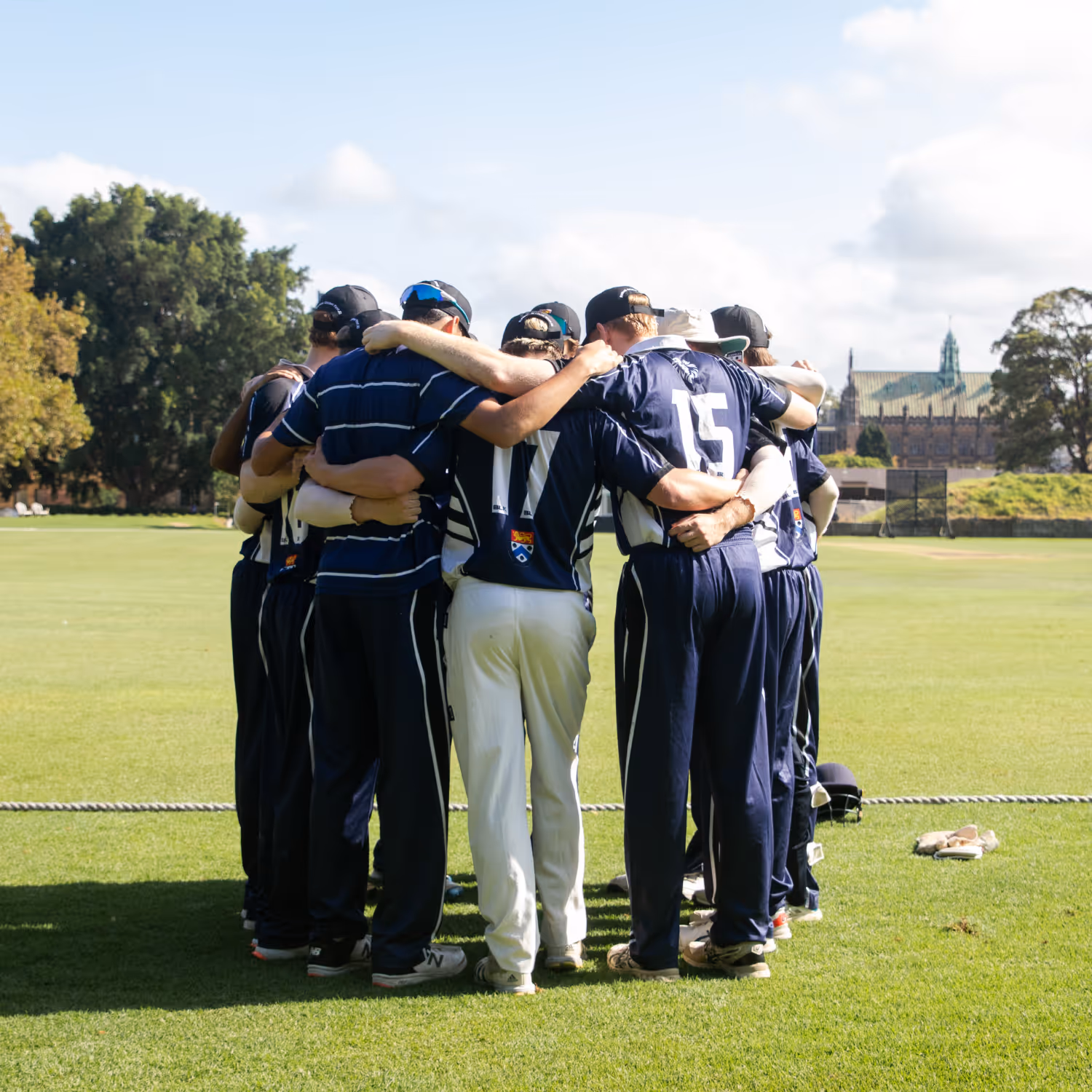 Group of cricket players in navy and white uniforms huddled together on a grassy field during daytime.