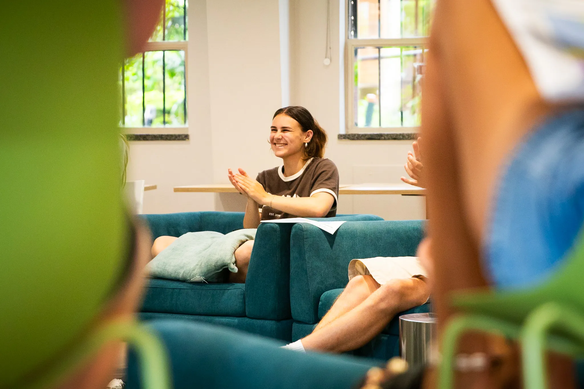 Young woman sitting on a teal couch clapping and smiling in a bright room with two windows.