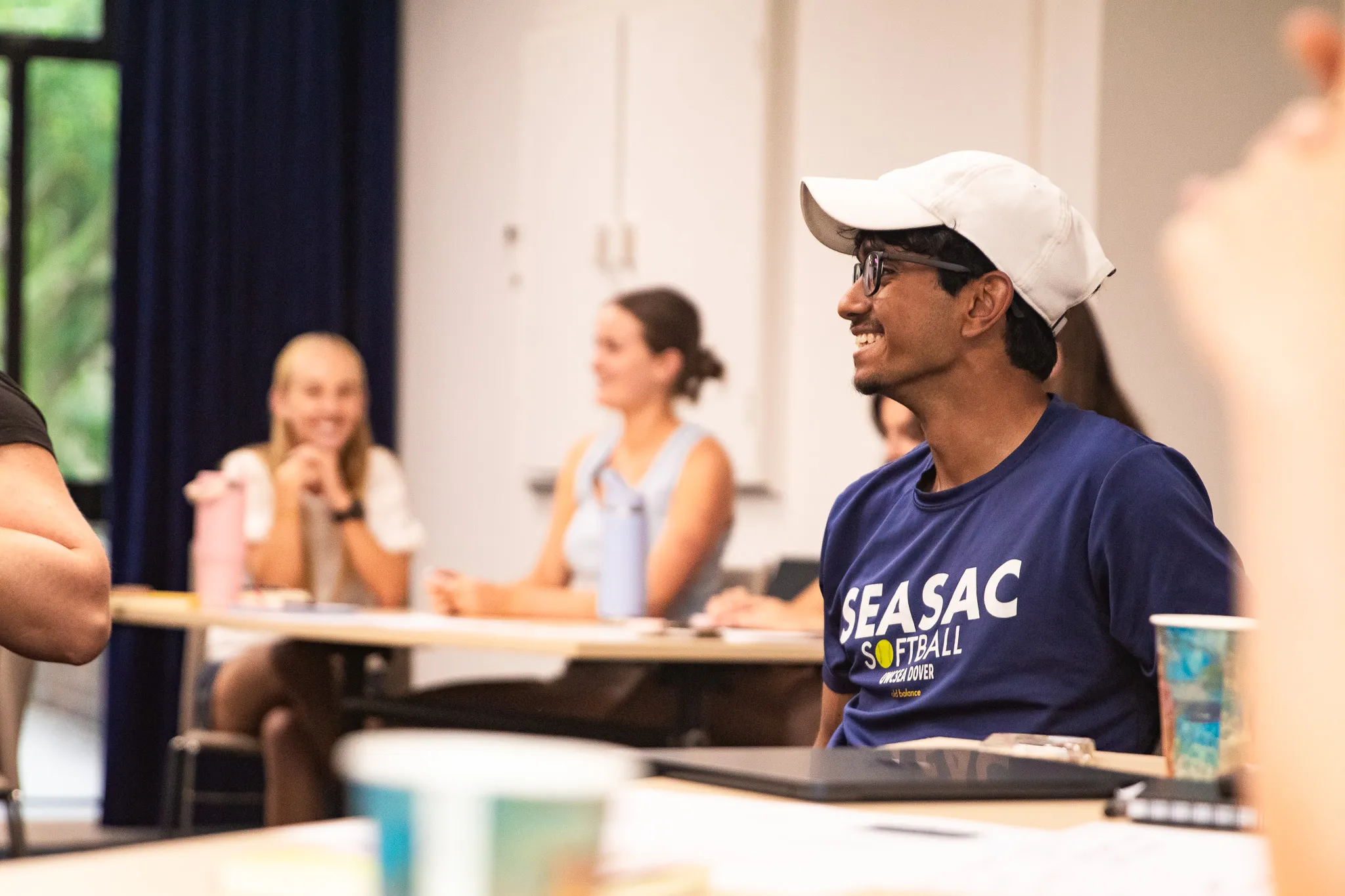 Young man in a white cap and glasses smiling while sitting in a classroom with other students.