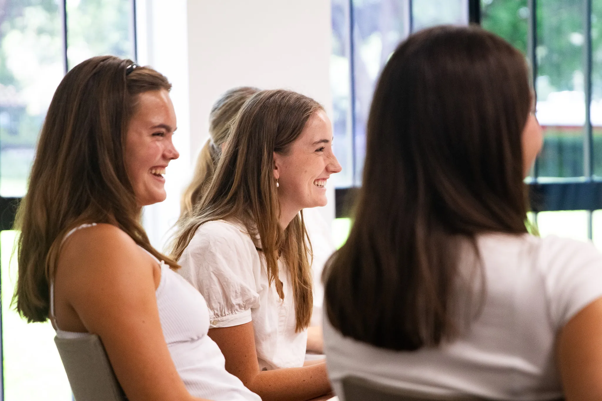 Three young women sitting and smiling indoors near large windows with greenery outside.