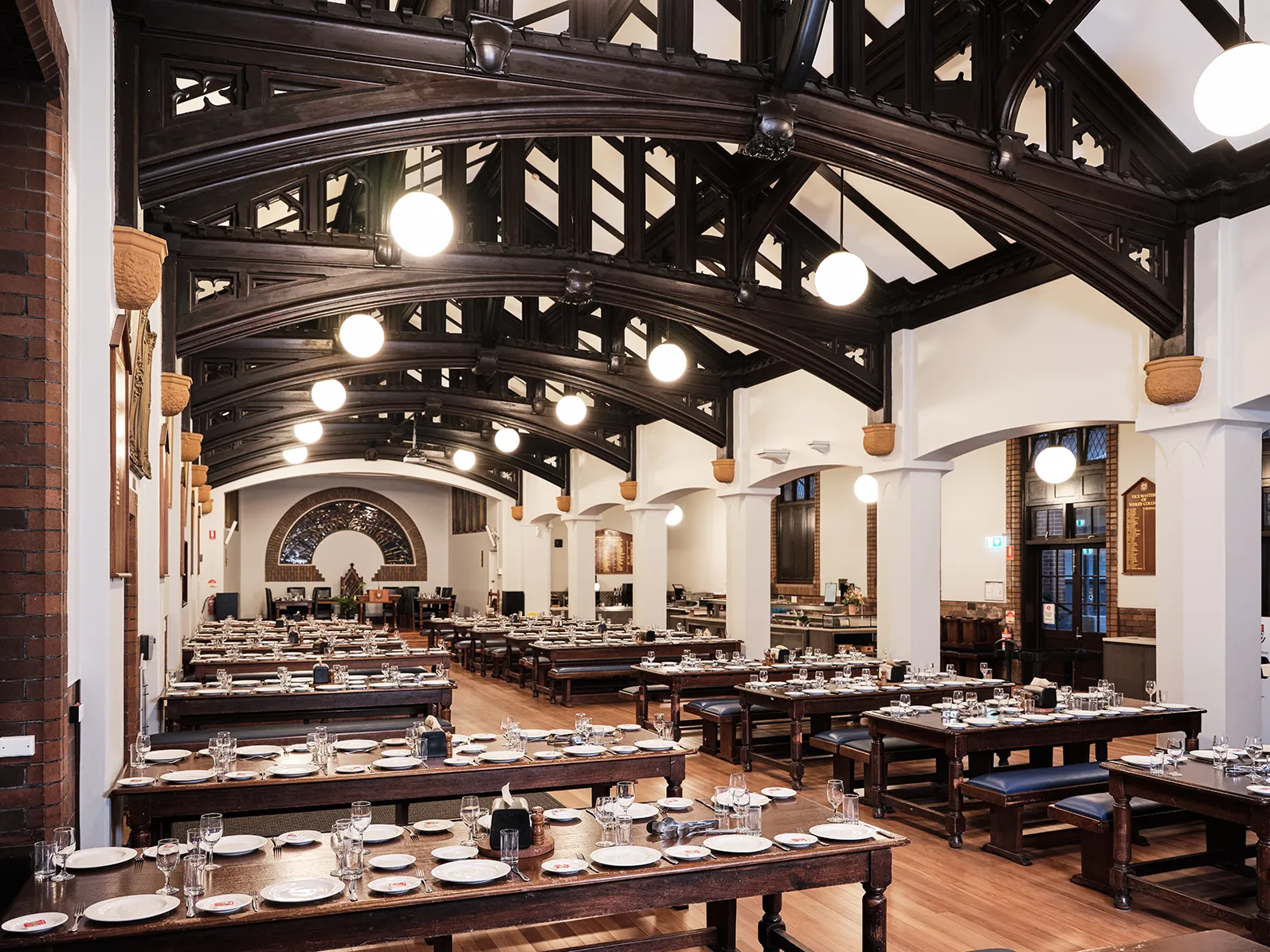 Large dining hall with wooden tables and benches set with plates and glasses under dark wooden arched beams and hanging globe lights.