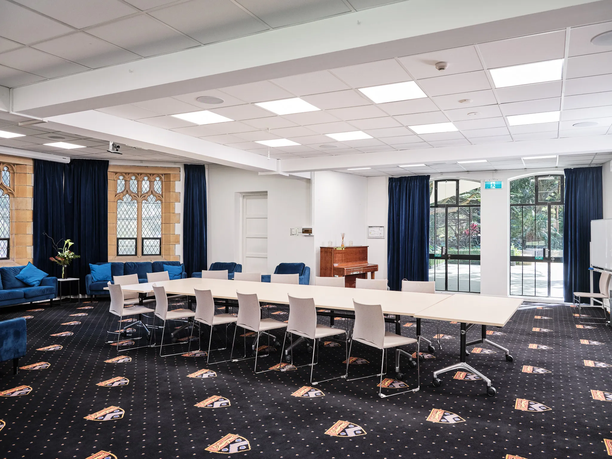 Spacious conference room with white tables arranged in a U shape surrounded by beige chairs, blue sofas, dark carpet with emblems, and large windows with blue curtains.