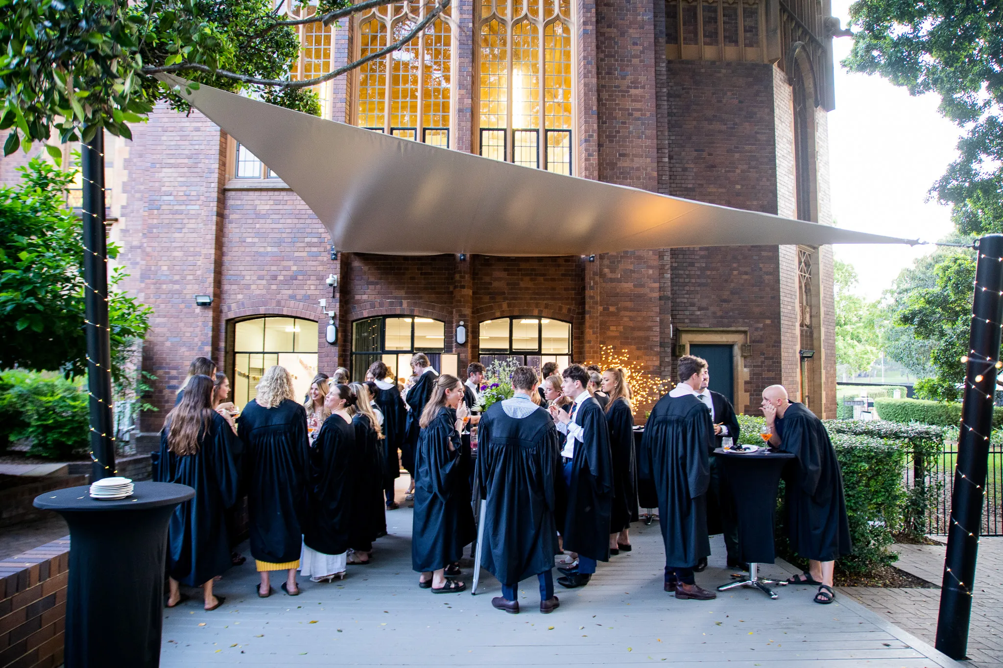 Group of people wearing black academic gowns socializing outdoors under a large triangular canopy in front of a brick building with tall windows.