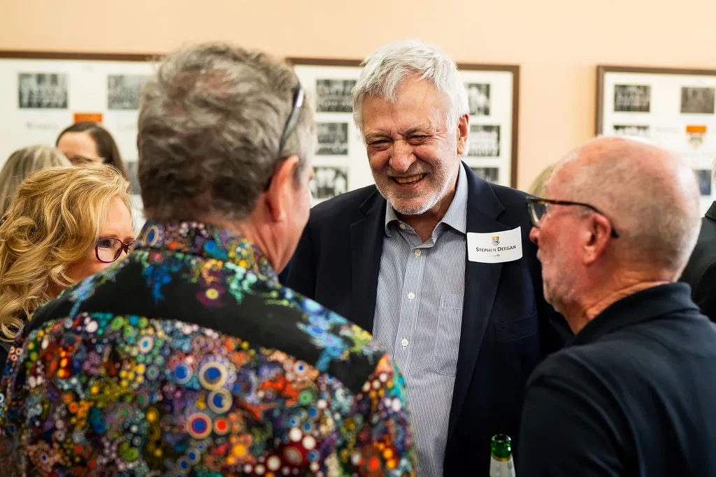 Smiling older man wearing a name tag engaged in conversation with two men and a woman at a social gathering.