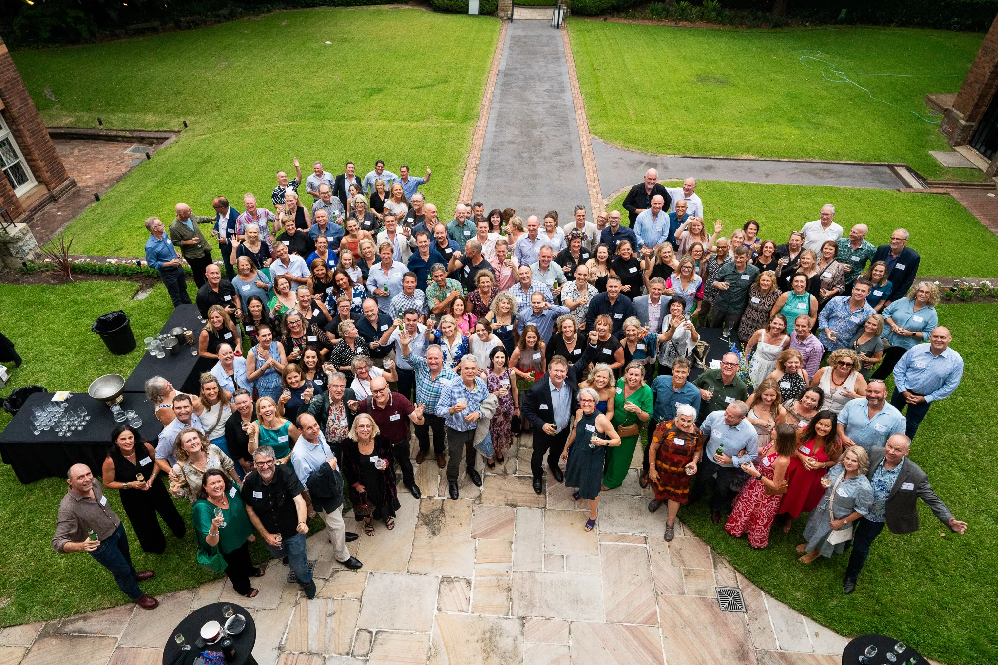 Large group of adults posing outdoors on a lawn and stone patio, many holding drinks and smiling at the camera.