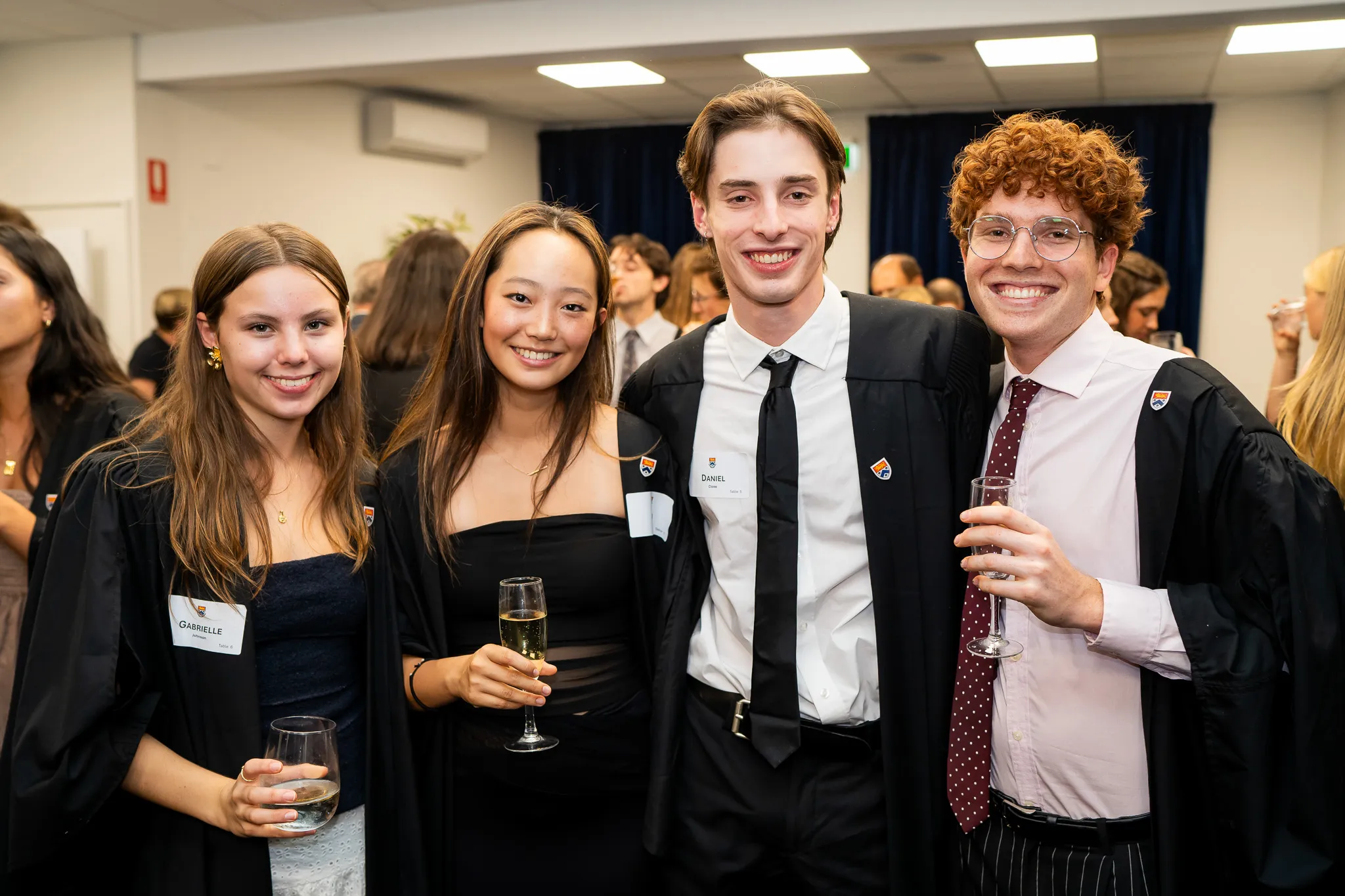 Four young adults in graduation gowns smiling and holding glasses at a celebration event.