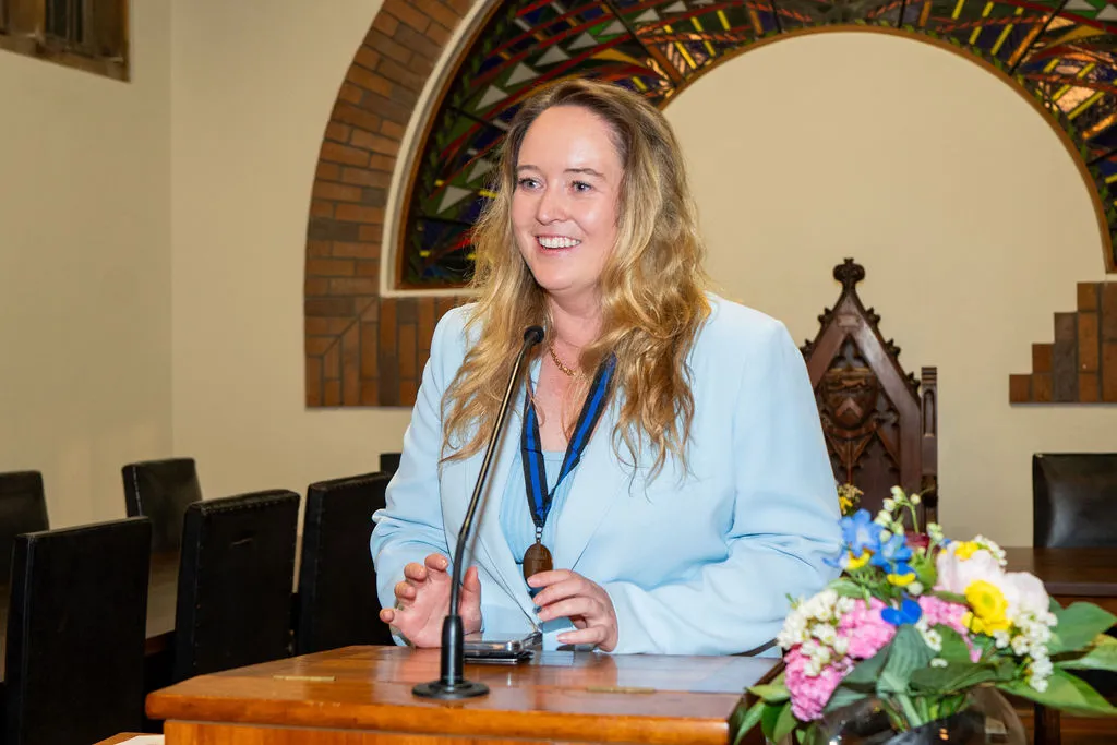 Smiling woman with long blonde hair wearing a light blue blazer speaking at a wooden podium with a microphone and floral arrangement, in a room with arched brick and stained glass windows.