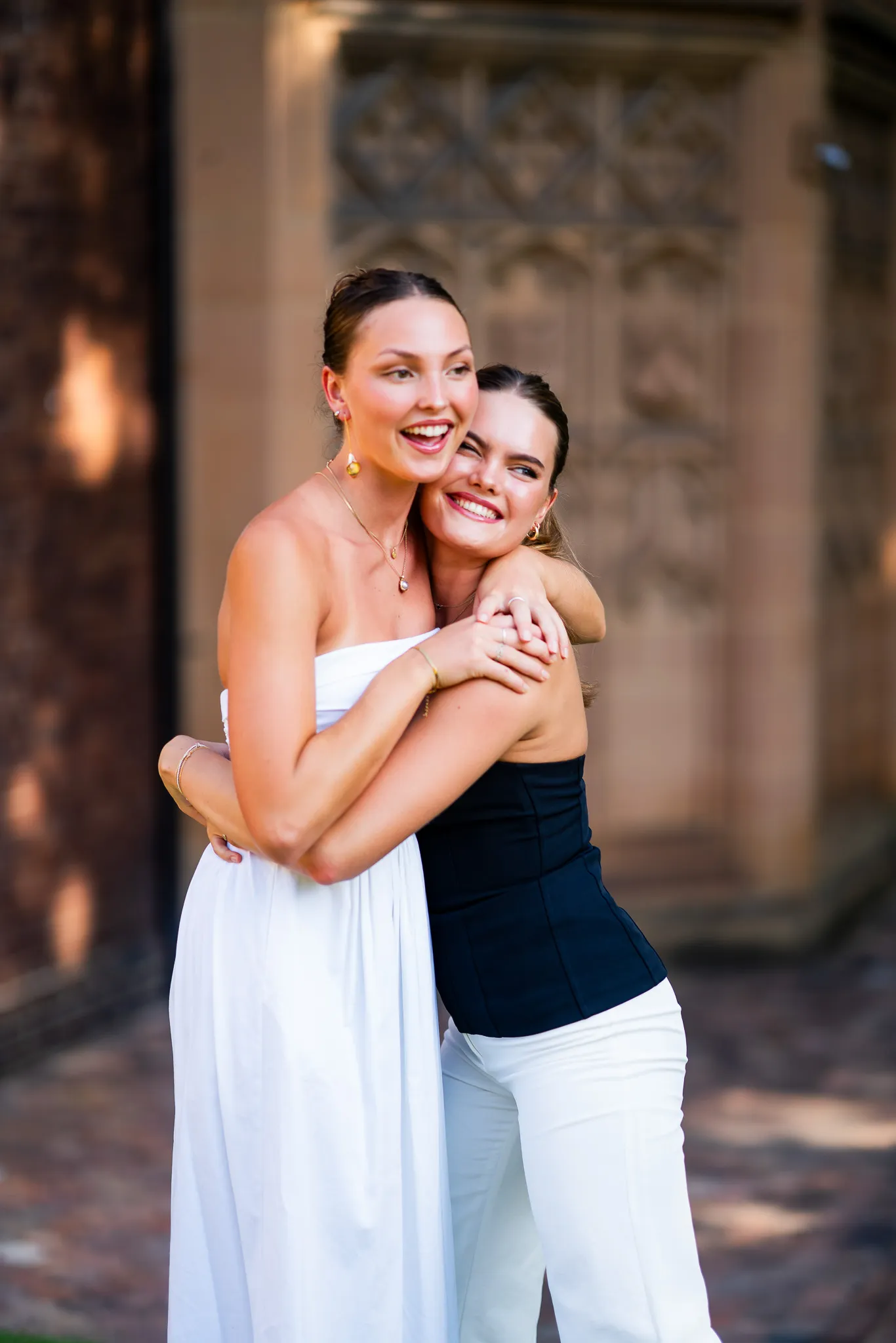 Two smiling women hugging outdoors, one wearing a white strapless dress and the other in a black strapless top with white pants.