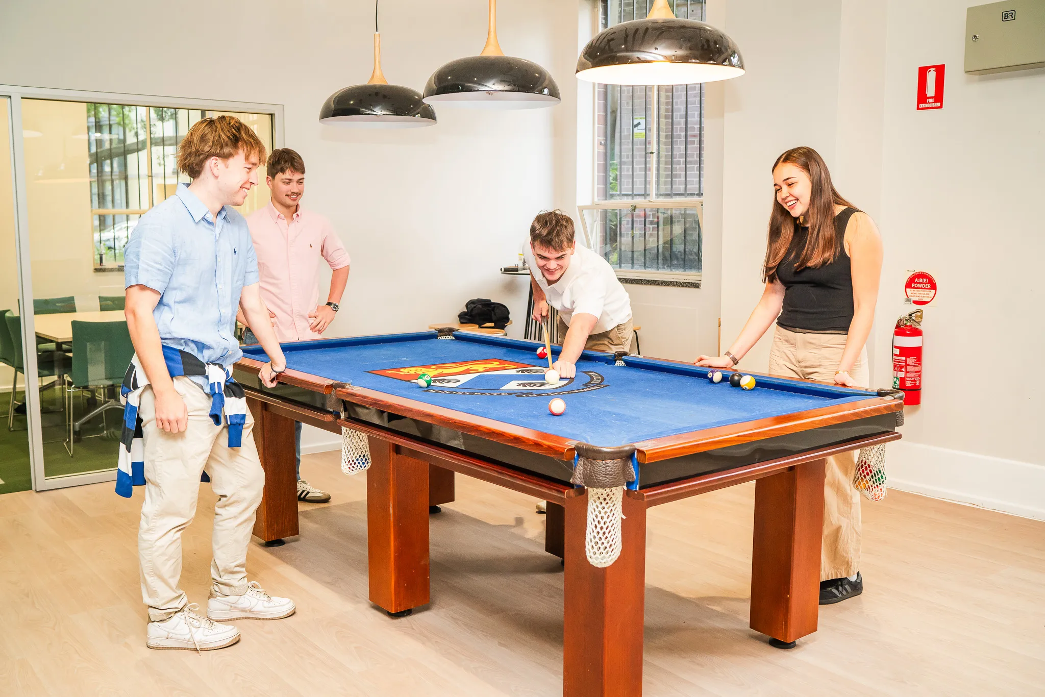 Four young adults smiling and playing pool on a blue-felt billiards table in a bright room.
