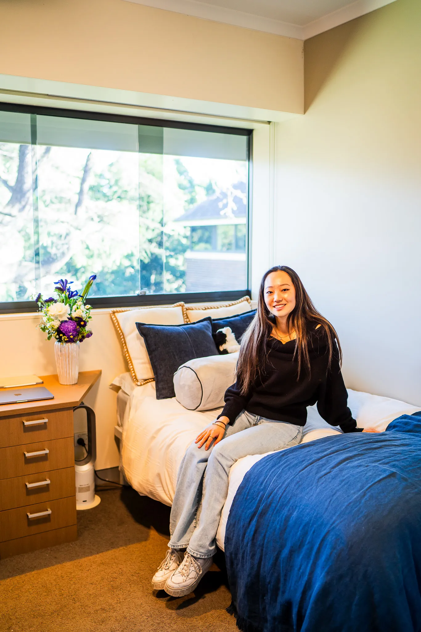 Smiling young woman sitting on the edge of a neatly made bed with blue and white bedding in a bright bedroom with a window view and wooden bedside table with flowers.