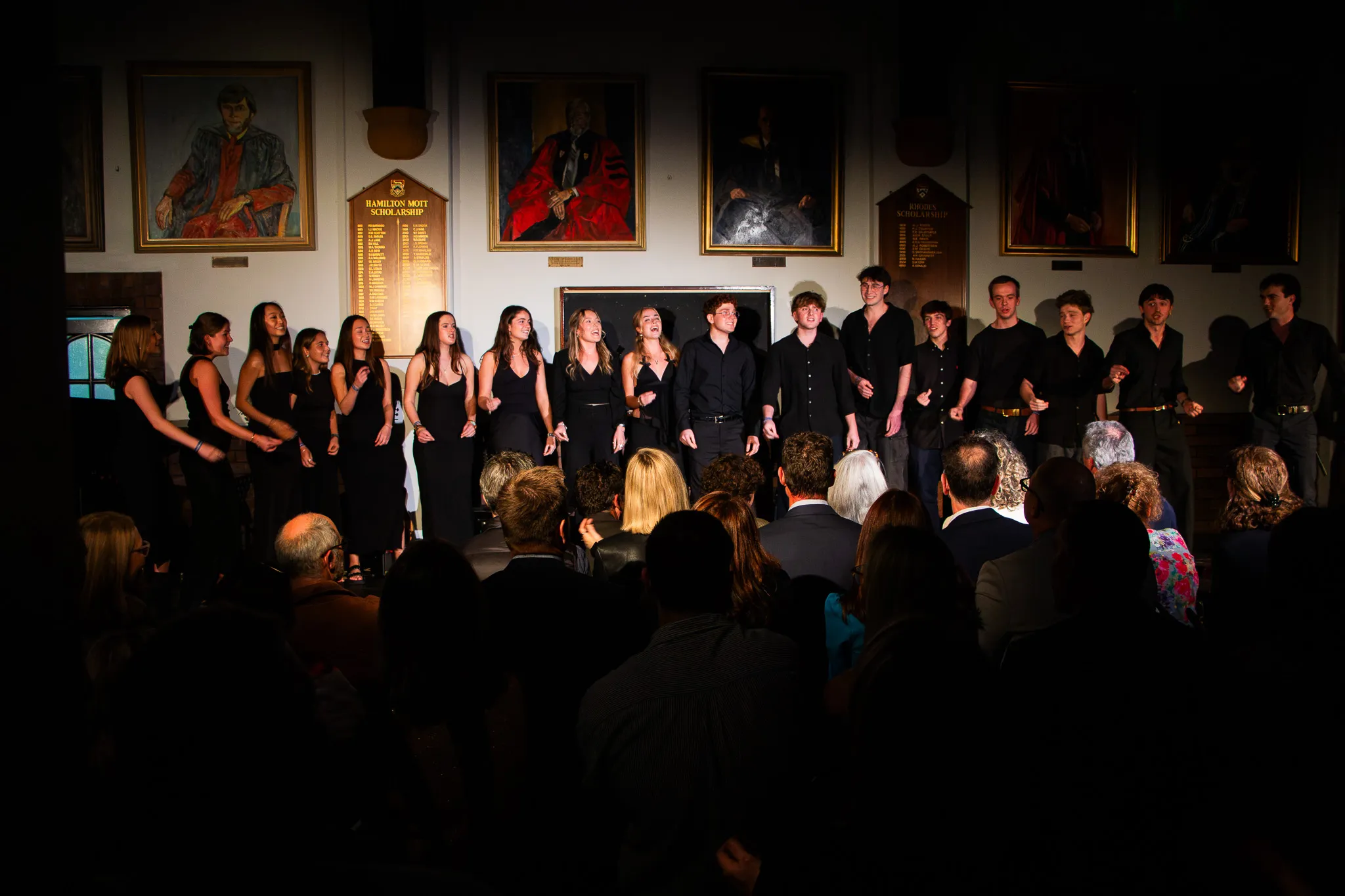 Group of young men and women dressed in black performing on stage in front of an audience in a room with portraits and scholarship plaques on the wall.