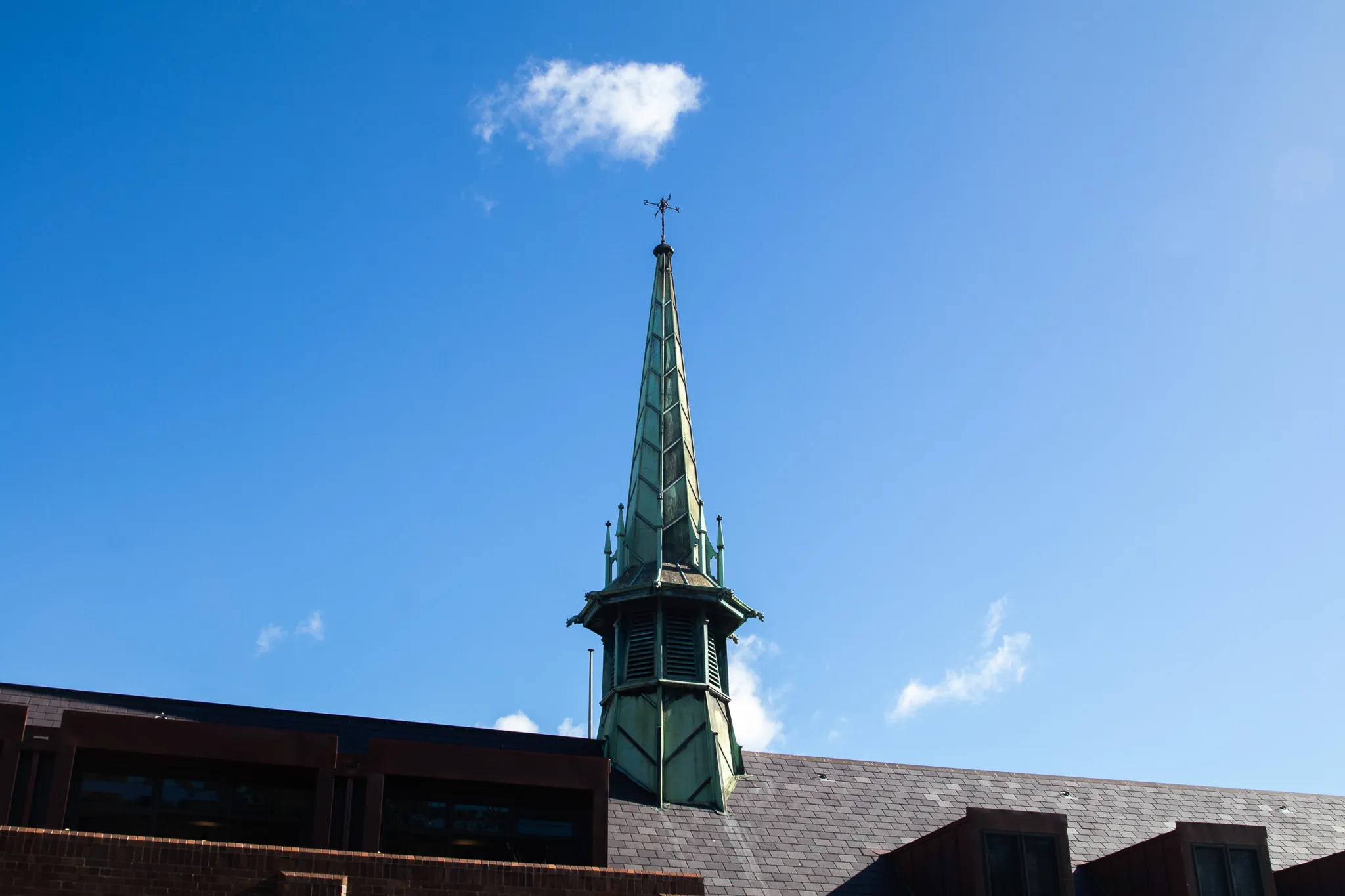 Tall steeple with a weather vane on a building roof under a clear blue sky with a few small clouds.
