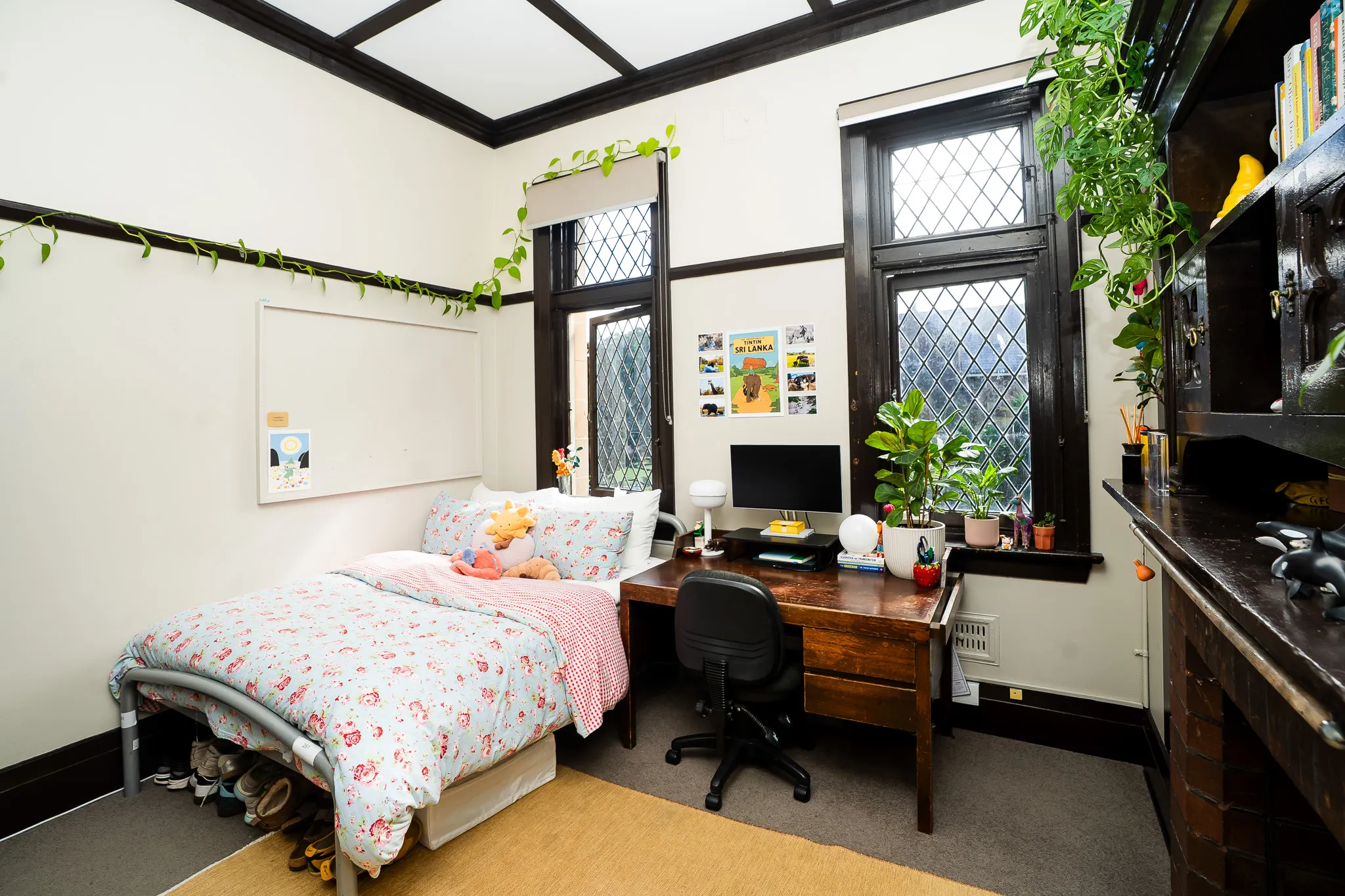 Cozy bedroom with floral bedding, a wooden desk with a computer, several plants, and diamond-patterned windows.