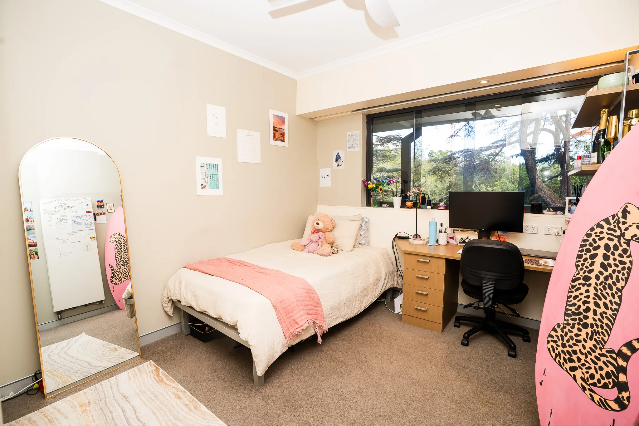 Cozy bedroom with a single bed adorned with a teddy bear and pink throw, a desk with a computer, a large window showing trees outside, and a standing mirror reflecting part of the room.