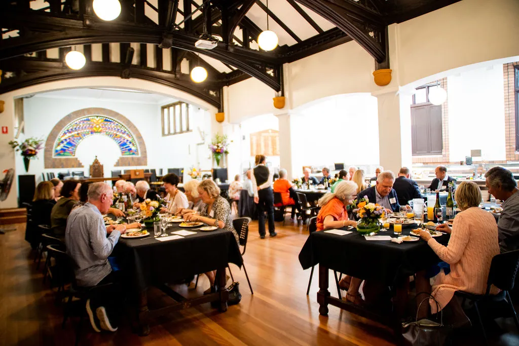 Group of people dining and conversing at tables with black tablecloths inside a well-lit room with wooden beams and stained glass window.