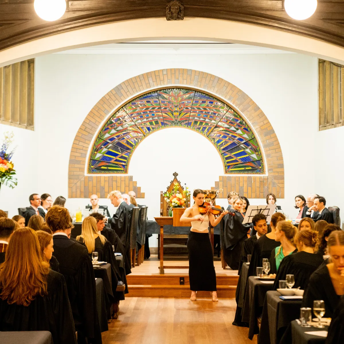 Woman playing violin in a formal dining hall with guests seated on both sides and a colorful stained glass arch in the background.