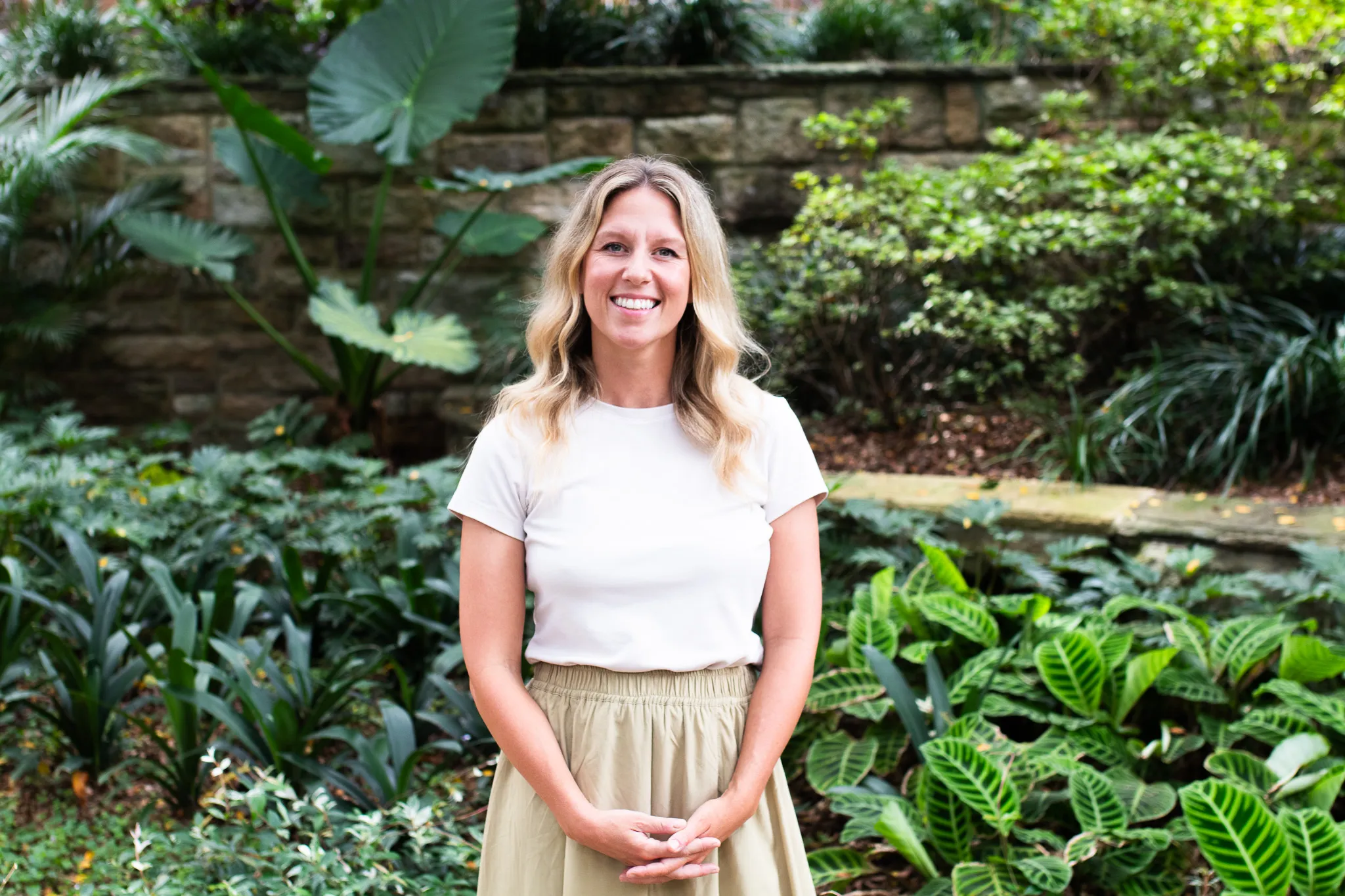 Smiling woman with wavy blonde hair wearing a white shirt and beige skirt standing in a lush garden.