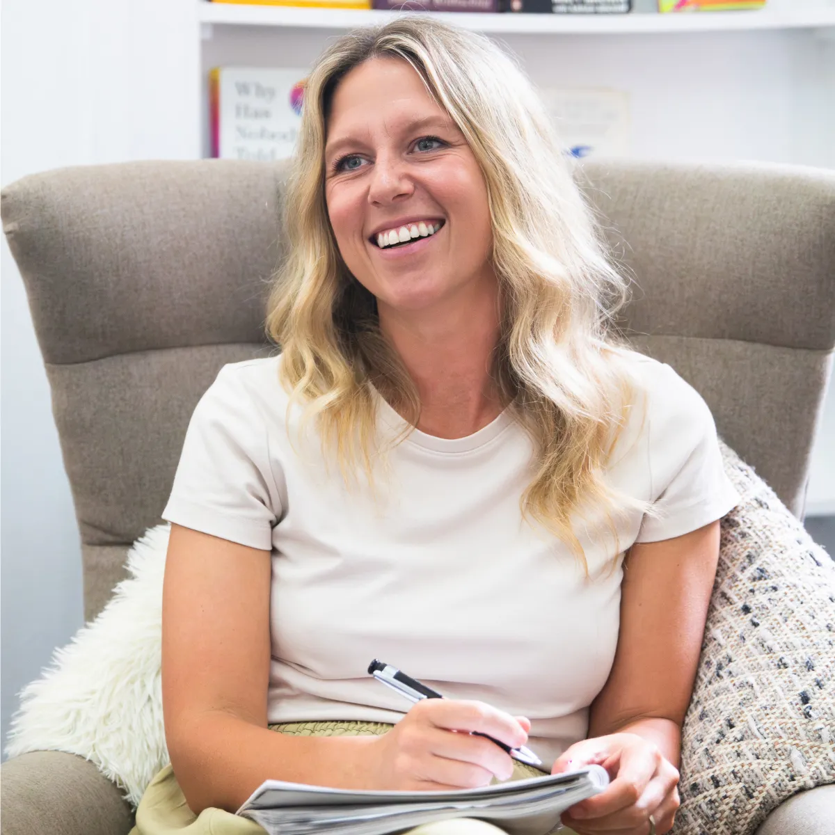 Smiling blonde woman sitting in a cushioned armchair, holding a pen and notebook.
