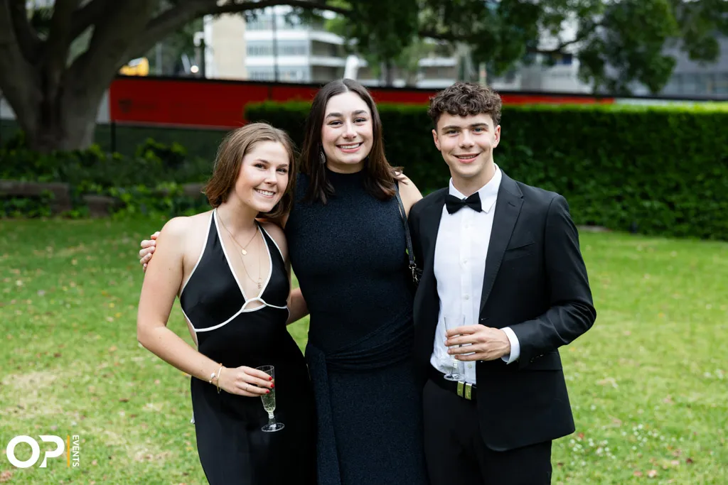 Three young adults dressed formally, smiling and posing outdoors on grass with drinks in hand.