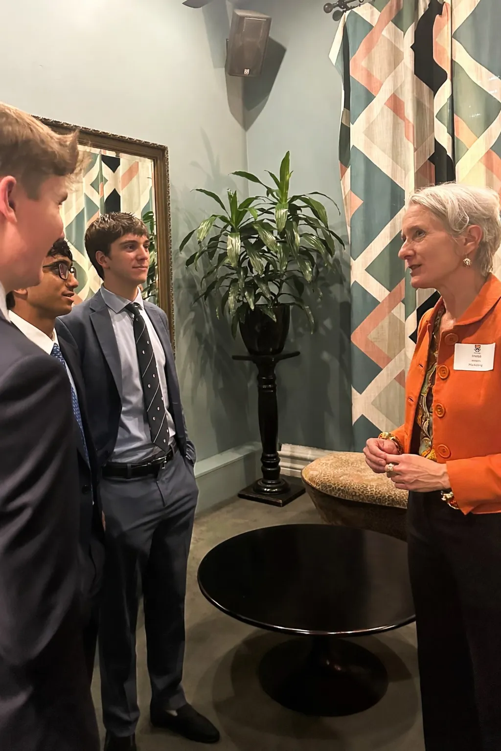 A woman in an orange jacket with a name tag is talking to three young men in suits and ties, standing in a room with a black round table, a potted plant on a stand, a large mirror, and patterned curtains.