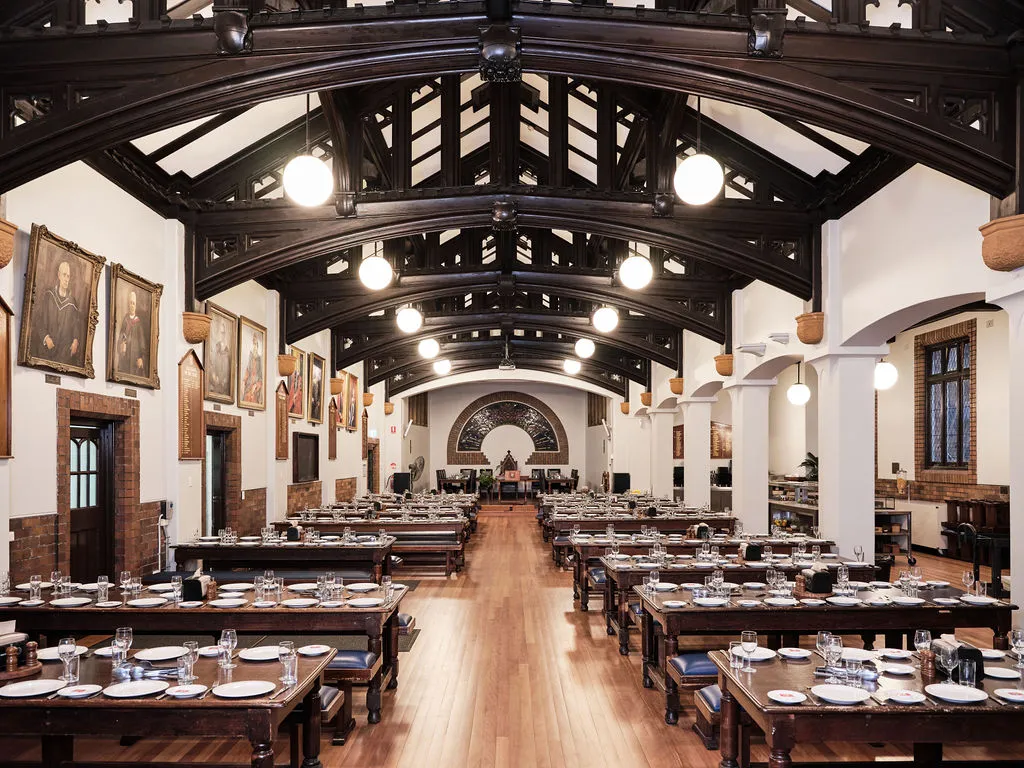 Spacious dining hall with wooden tables and benches neatly set with plates and glasses under a high, dark wooden beam ceiling and framed portraits on white walls.