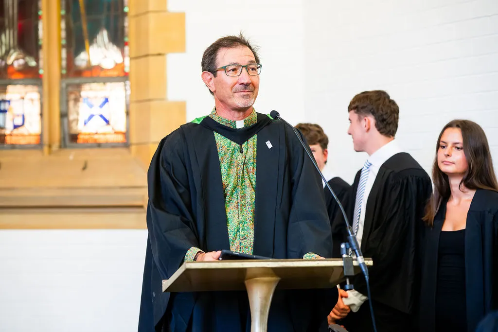 Man wearing academic gown and colorful shirt standing at podium with microphone during a ceremony, with three young adults in gowns behind him.